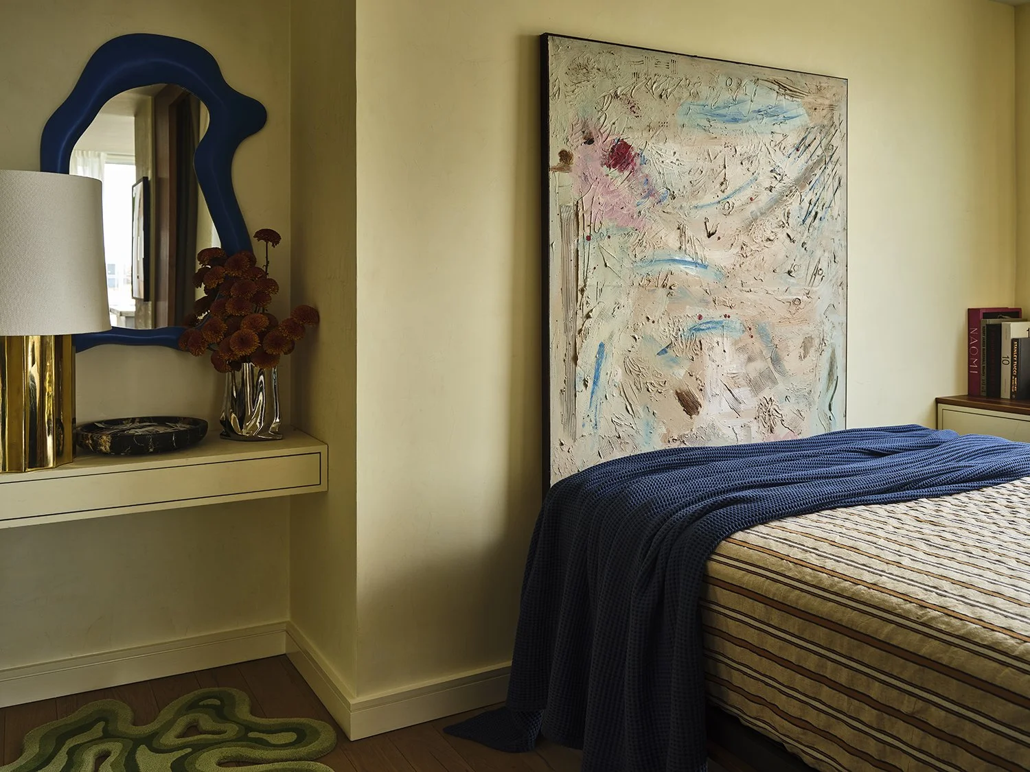 A bedroom with beige walls, a large abstract painting above the bed, a nightstand with books, and a mirror reflecting a window with curtains.