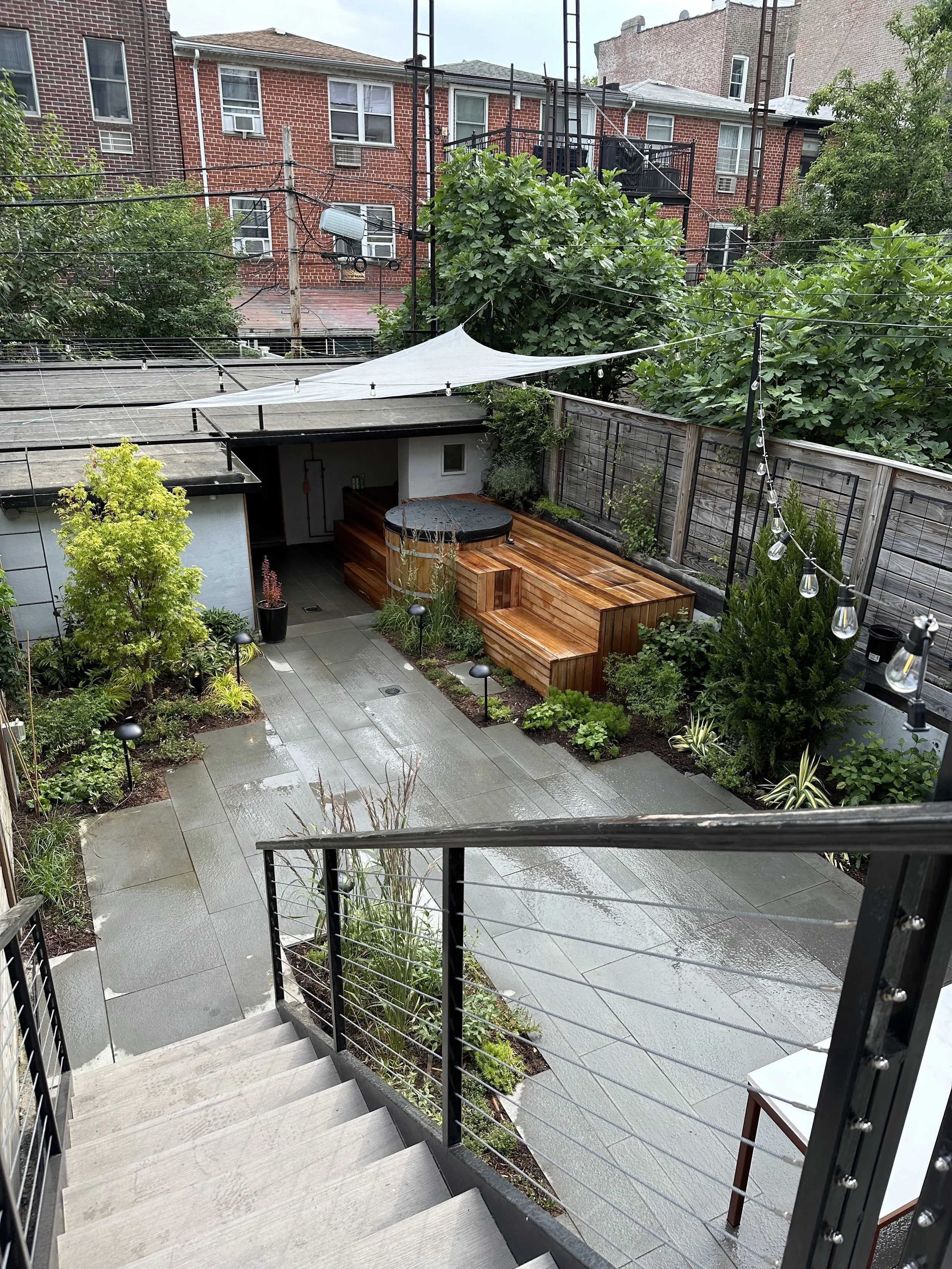 View of a backyard garden with wet concrete patio, small trees, plants, and outdoor lighting, photograph taken from top of stairs.