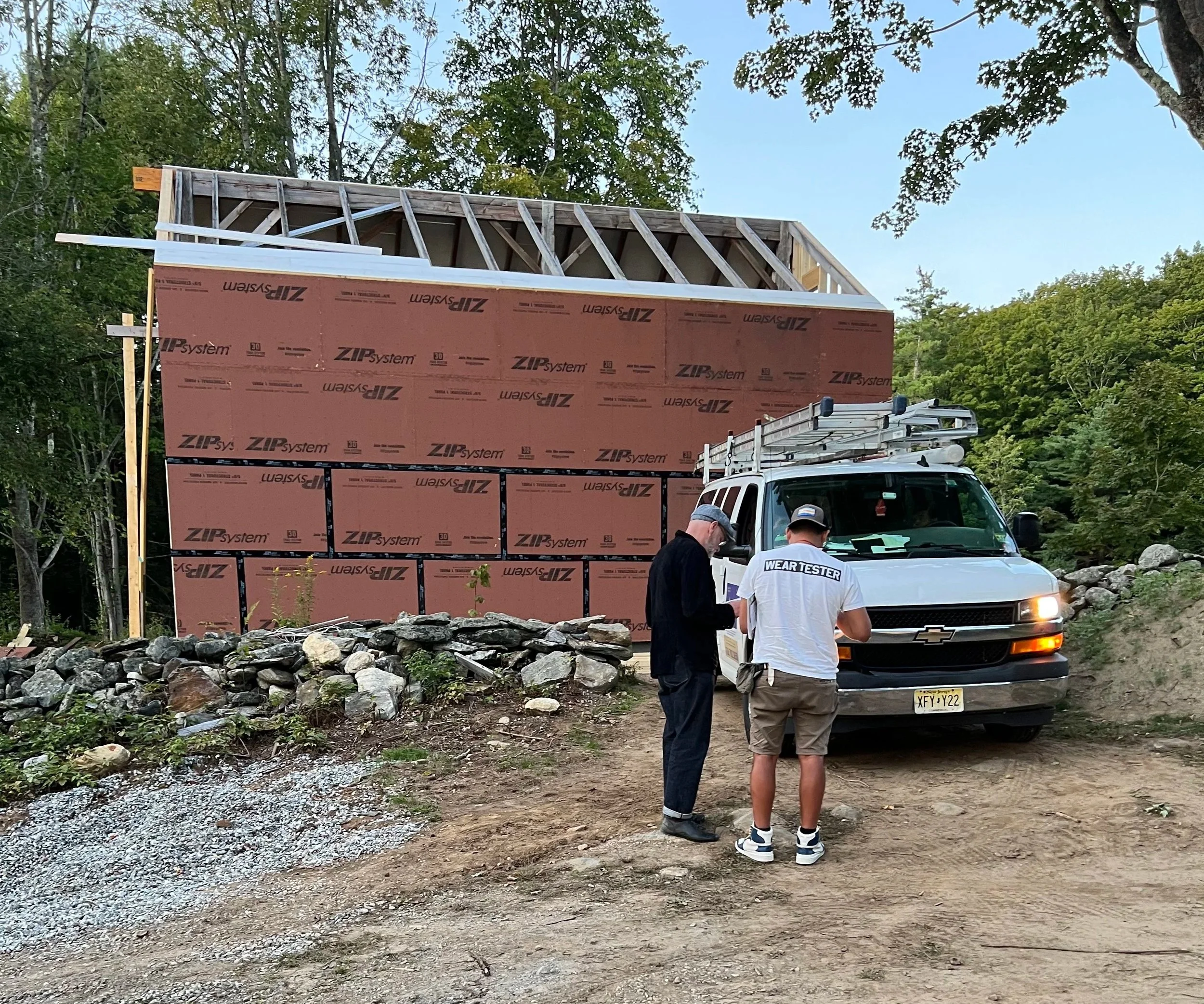 Two men standing in front of a white utility vehicle near a partially constructed building with pink insulation panels, surrounded by trees and rocks.