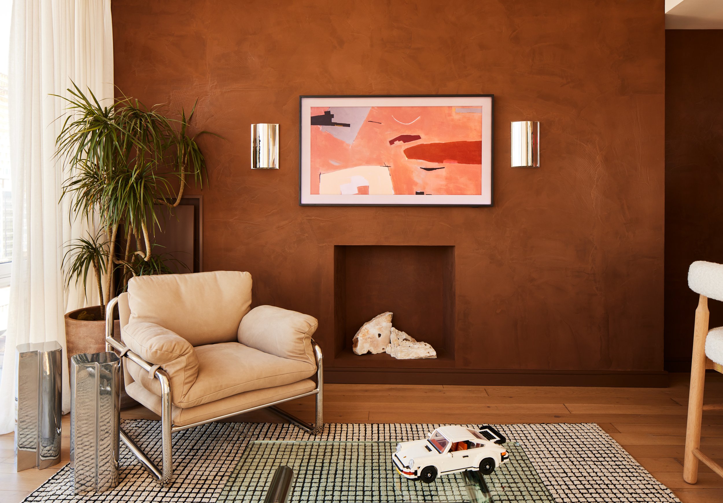 A modern living room with brown textured wall, abstract artwork, beige armchair, indoor plant, decorative items, and a white toy car on a patterned rug.