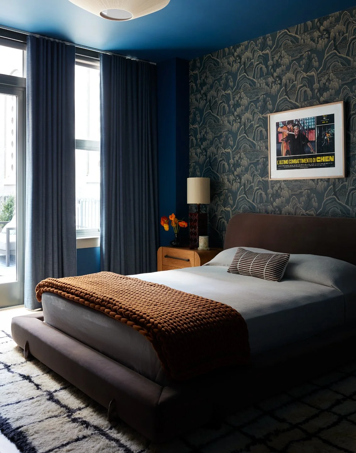 A bedroom with a bed, dark brown headboard, white bedding, and a brown knitted blanket. There is a wooden nightstand with a lamp and a vase with orange flowers. Blue curtains cover a window, and a framed movie poster is on the wall. The ceiling is blue, and there's a patterned black and white rug on the floor.