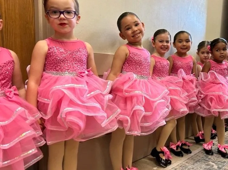 A group of young girls in matching pink ballet costumes with frilly skirts and black shoes with pink bows, standing in a line.