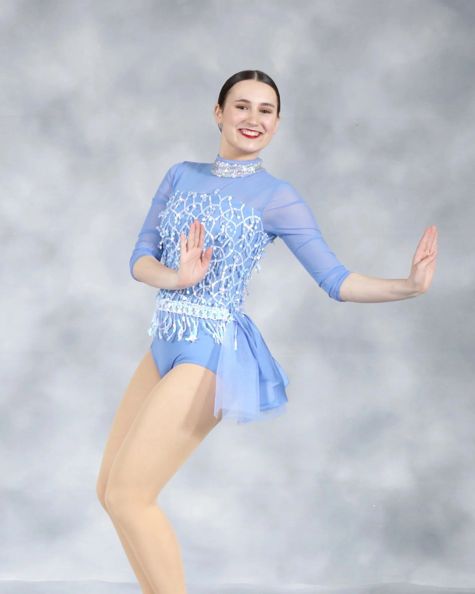 A woman in a blue figure skating dress and beige tights poses against a gray backdrop.