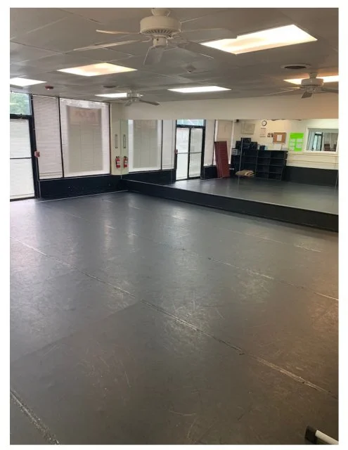 Empty dance studio with black flooring, large mirrors, and windows.