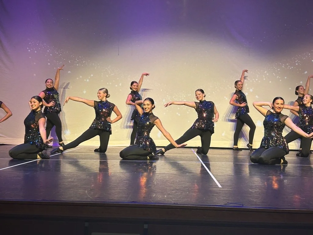 A group of female dancers on stage performing a dance routine. They are dressed in black, sparkly outfits and are striking various poses.