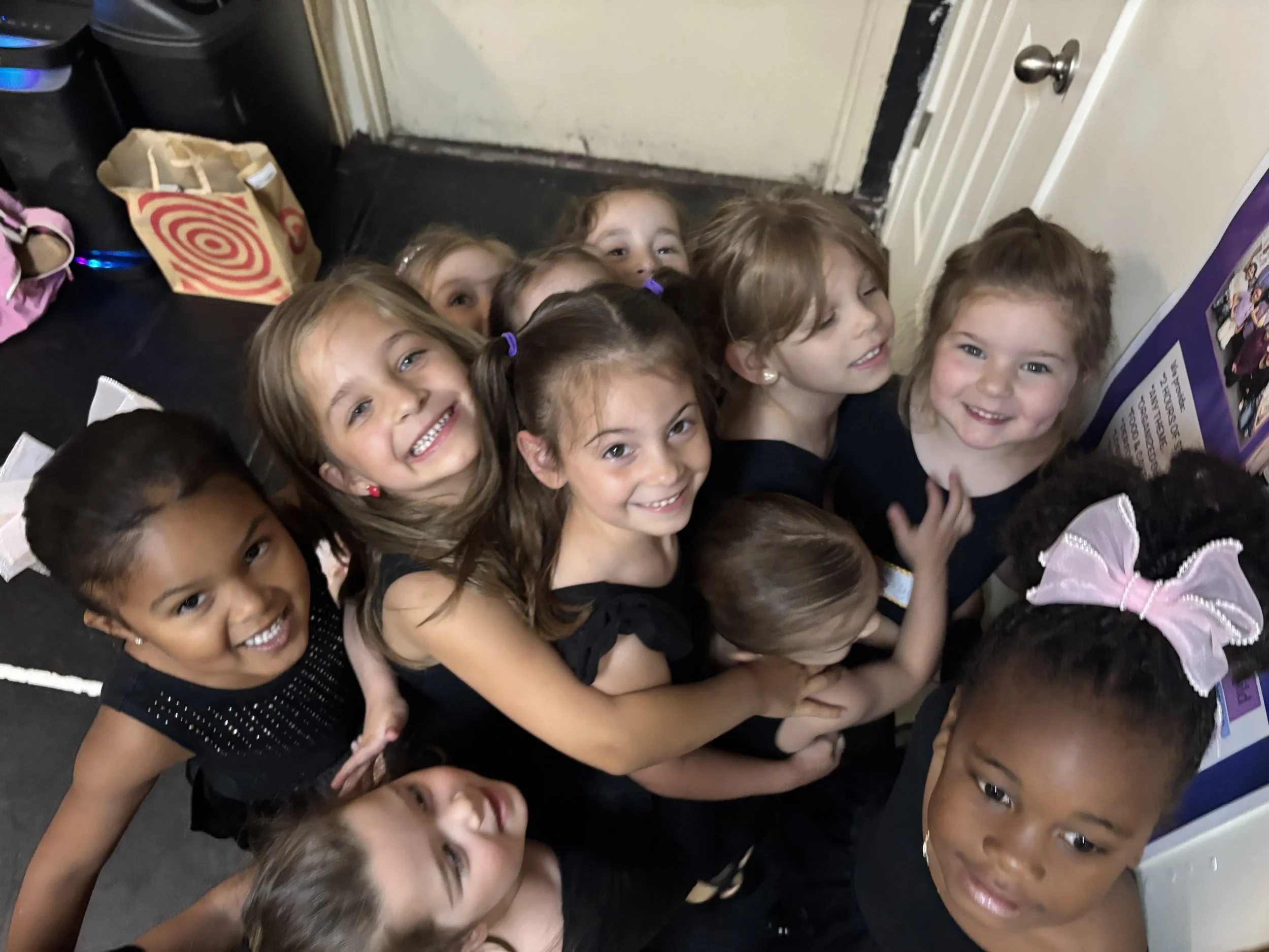 Group of smiling young girls standing close together indoors, near a door and a bulletin board.