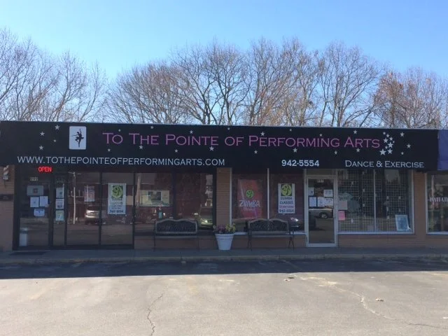 Storefront with signs advertising dance and exercise classes, with trees in the background and a parking lot in front.