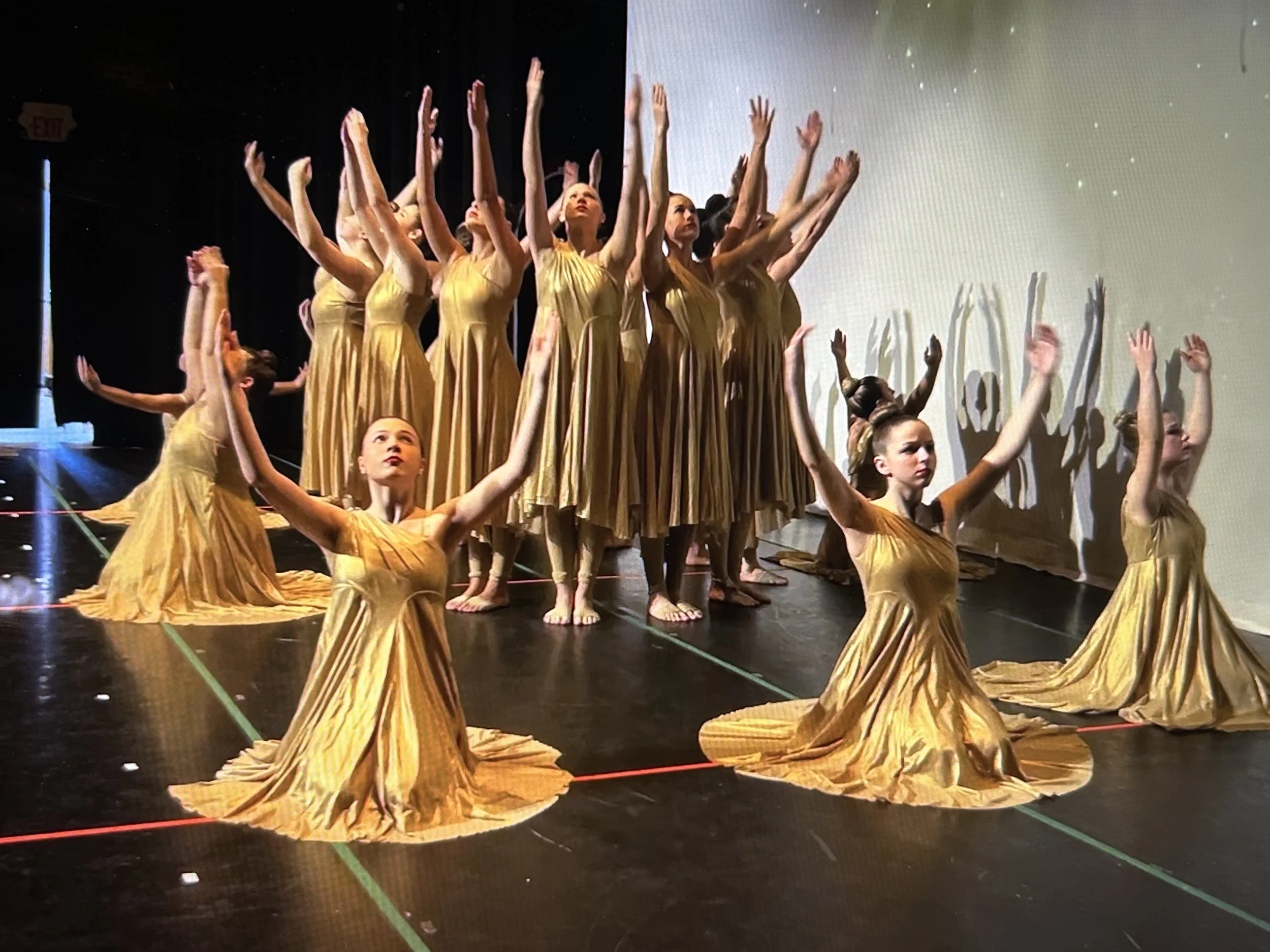 Dance performance with dancers in gold dresses, some kneeling, others standing with arms raised, casting shadows on the wall behind.