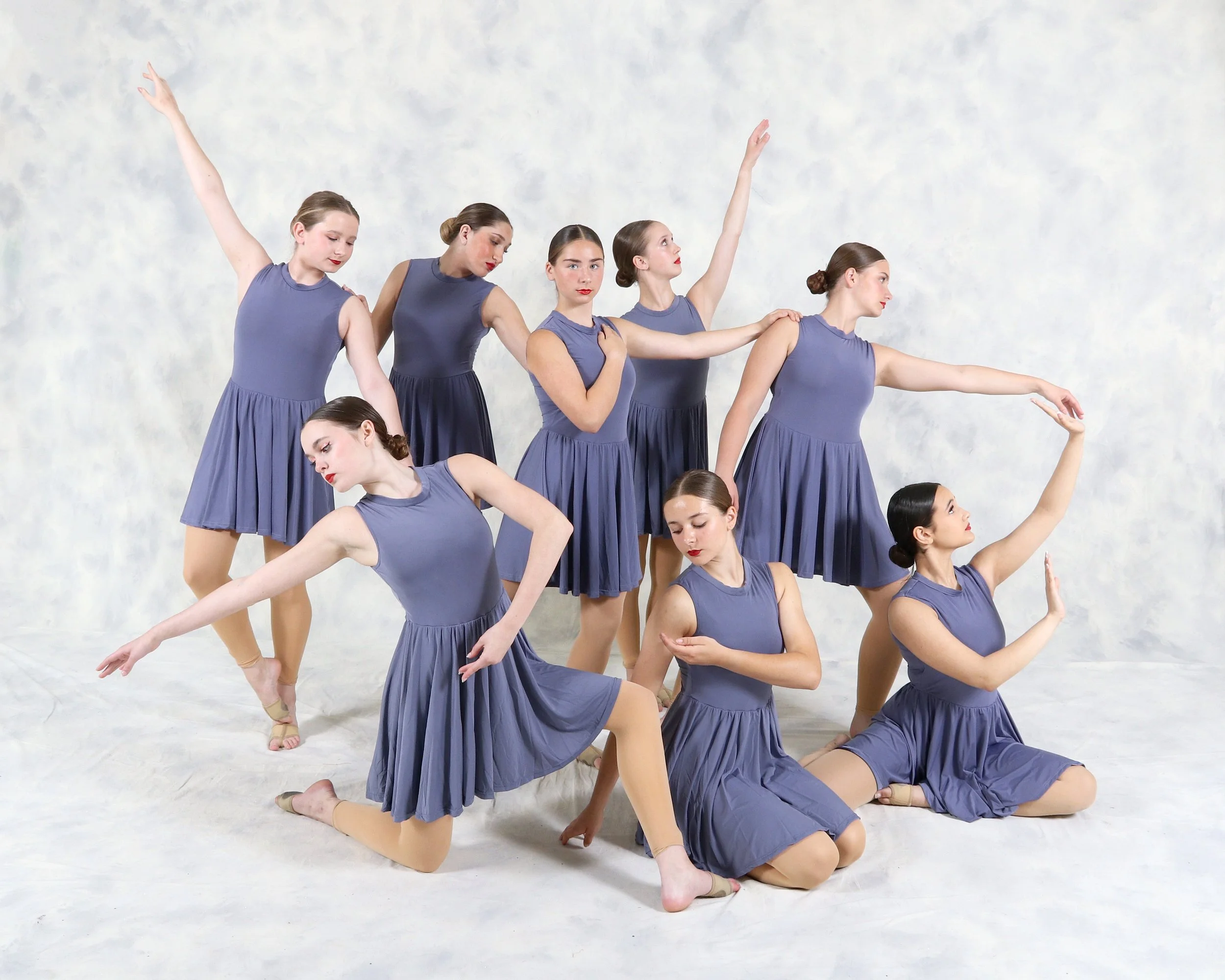 Eight young women in matching blue dresses practicing ballet poses on a white backdrop.