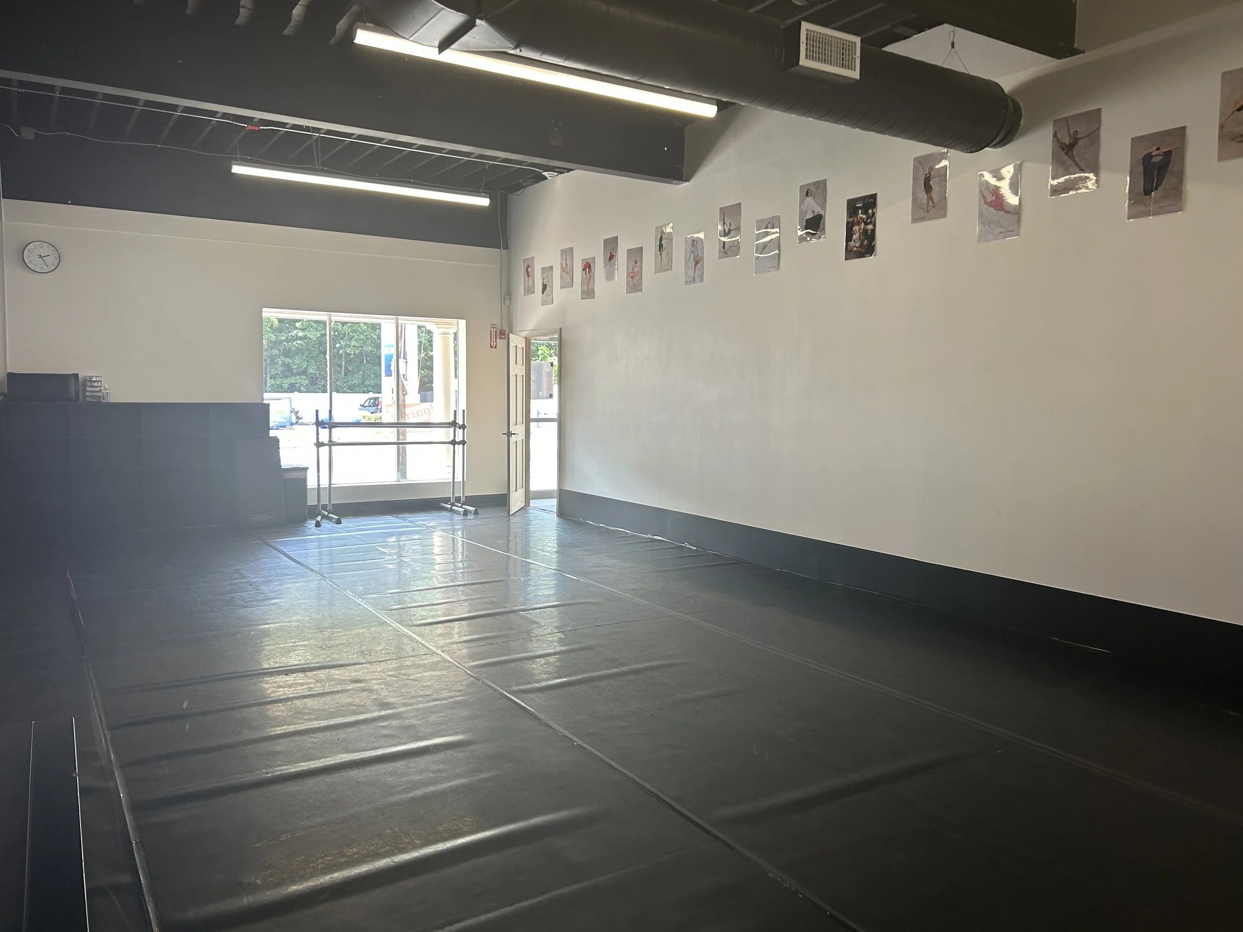 Interior of an empty dance studio with large windows, gray tiled floor, white walls, and a display of dance photos. The ceiling has exposed ductwork and lighting, and there are ballet barres near the windows.