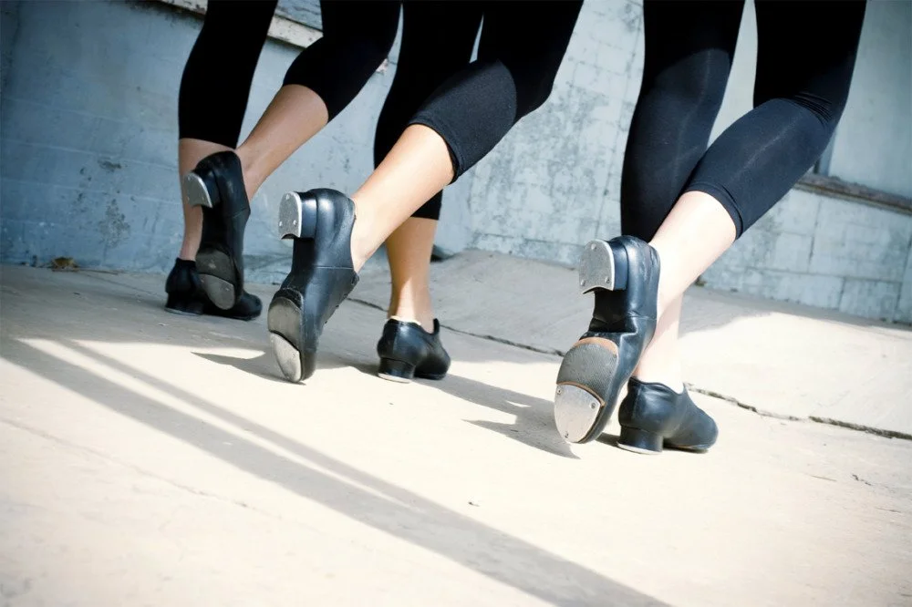 Two pairs of dancers in black shoes and black leggings practicing ballet dance on a concrete floor.