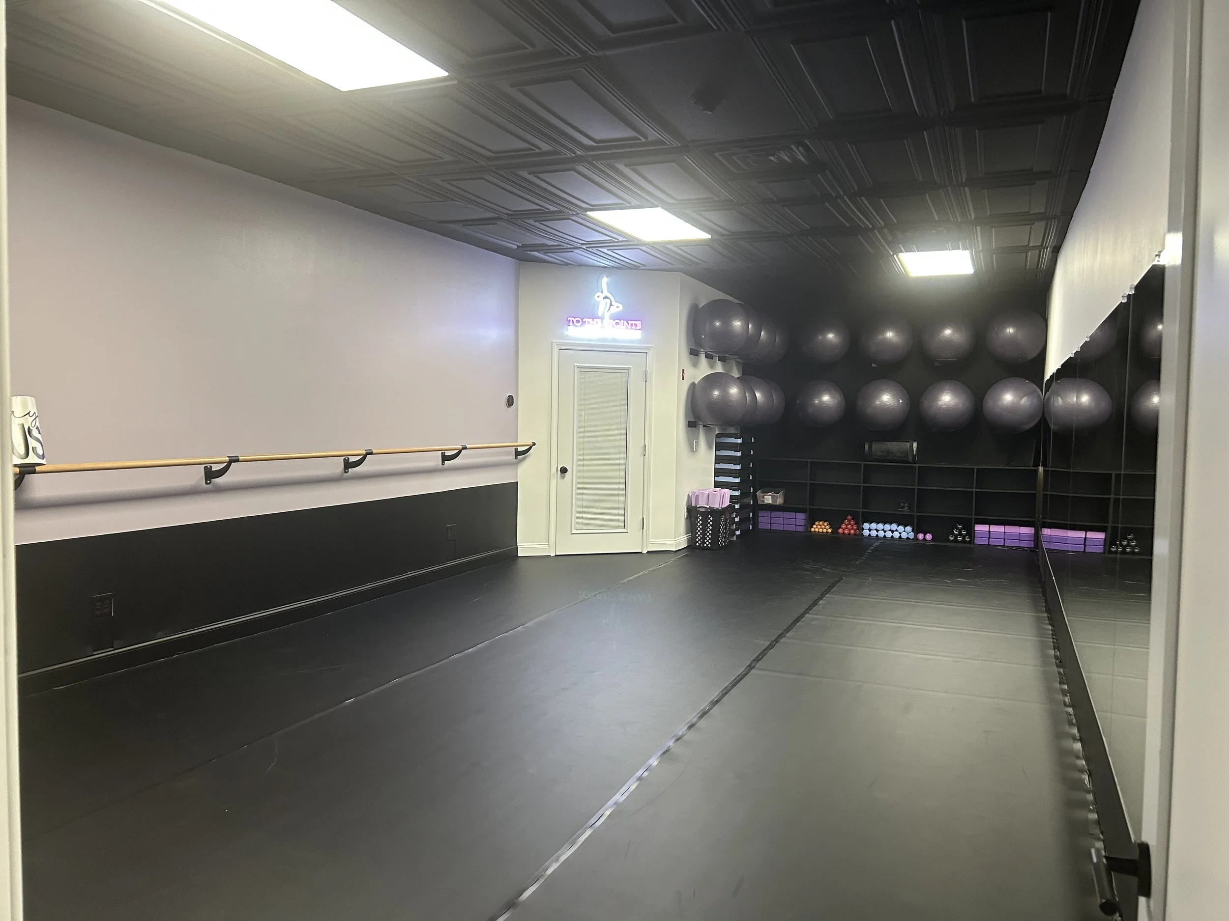Interior view of a dance or fitness studio with black flooring, a ballet barre on the left wall, a wall-mounted mirror on the right, and a corner with black storage shelves holding various exercise balls and equipment. The ceiling has a black, textured pattern with bright fluorescent lights, and there is a neon sign in the back corner next to a door.