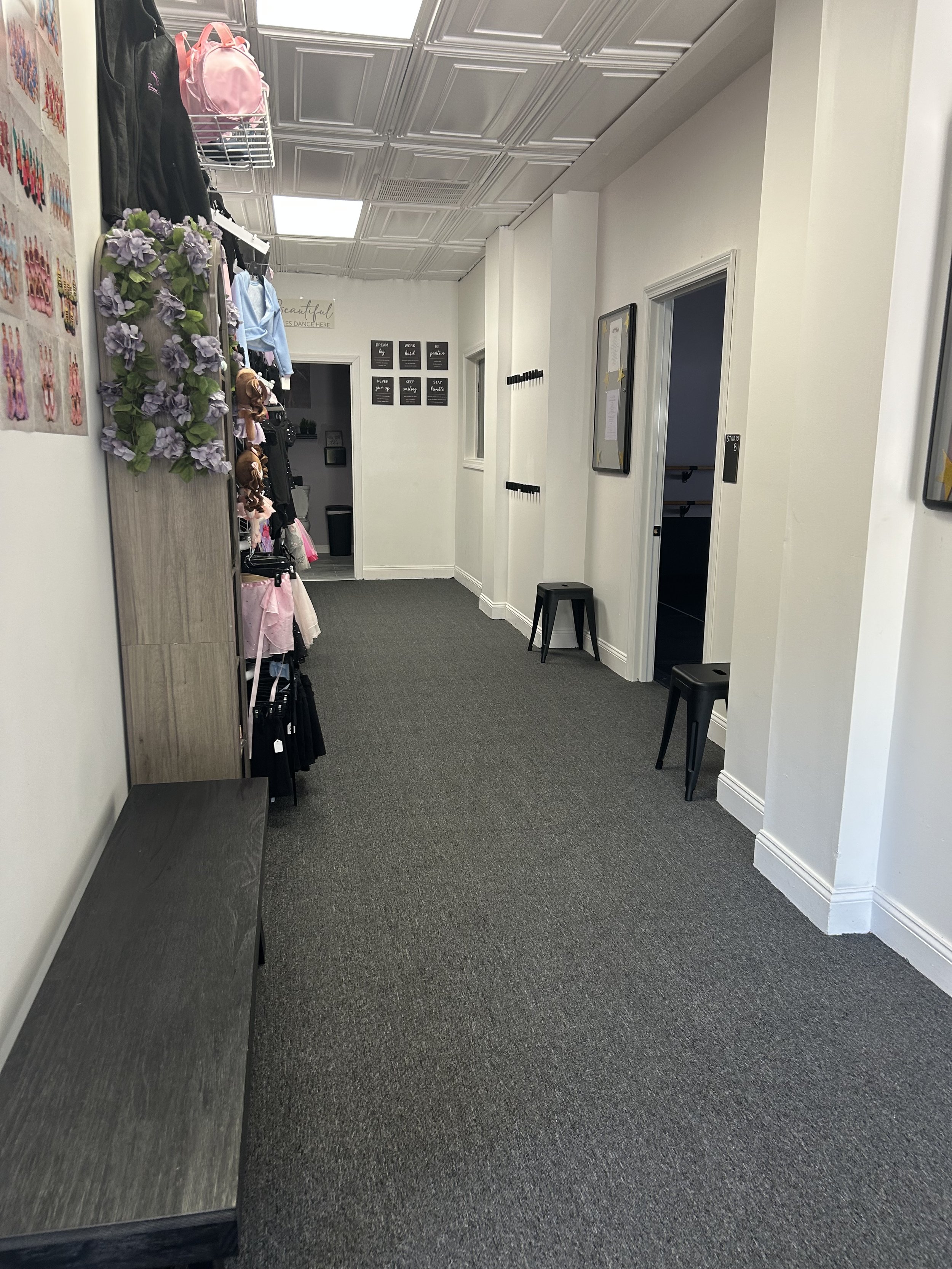 Hallway with white walls, gray carpet, black stools, and a door leading to a studio. There is a display of handbags and accessories on the left wall and motivational posters on the back wall.