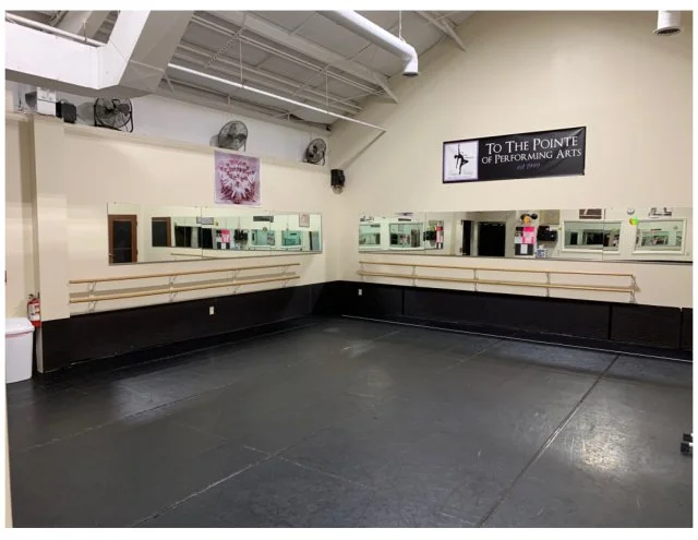 Dance studio with black floor, wall-mounted ballet barres, mirrors, and a sign that reads 'To The Pointe of Performing Arts'.