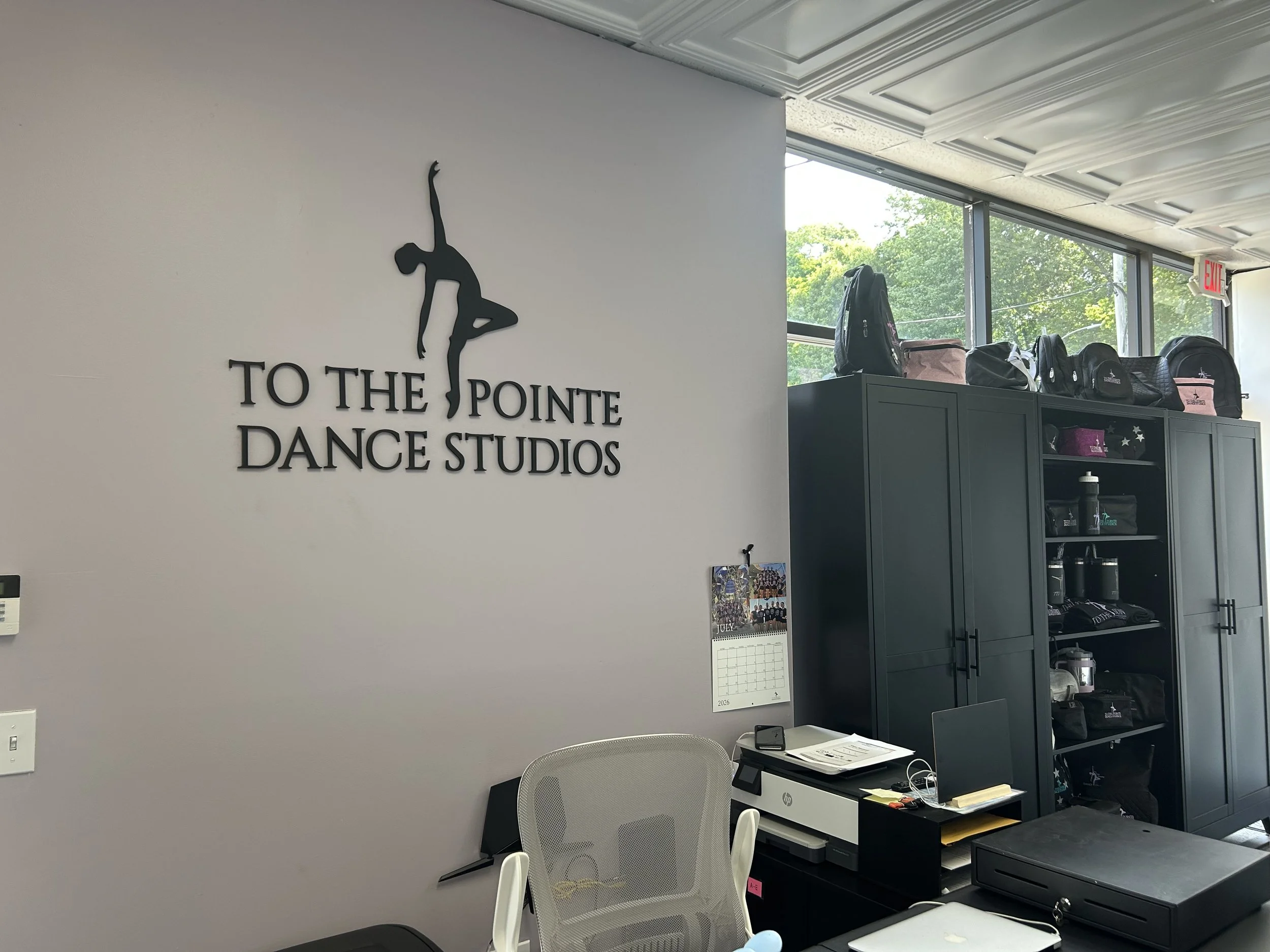 storefront with a sign that reads 'To The Pointe of Performing Arts' and a website URL, with large windows showing posters inside and two benches outside, with leafless trees in the background.