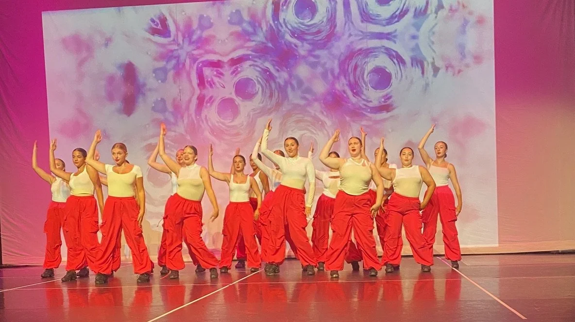Group of female dancers performing on stage, wearing white tops and red wide-legged pants with a colorful abstract backdrop.