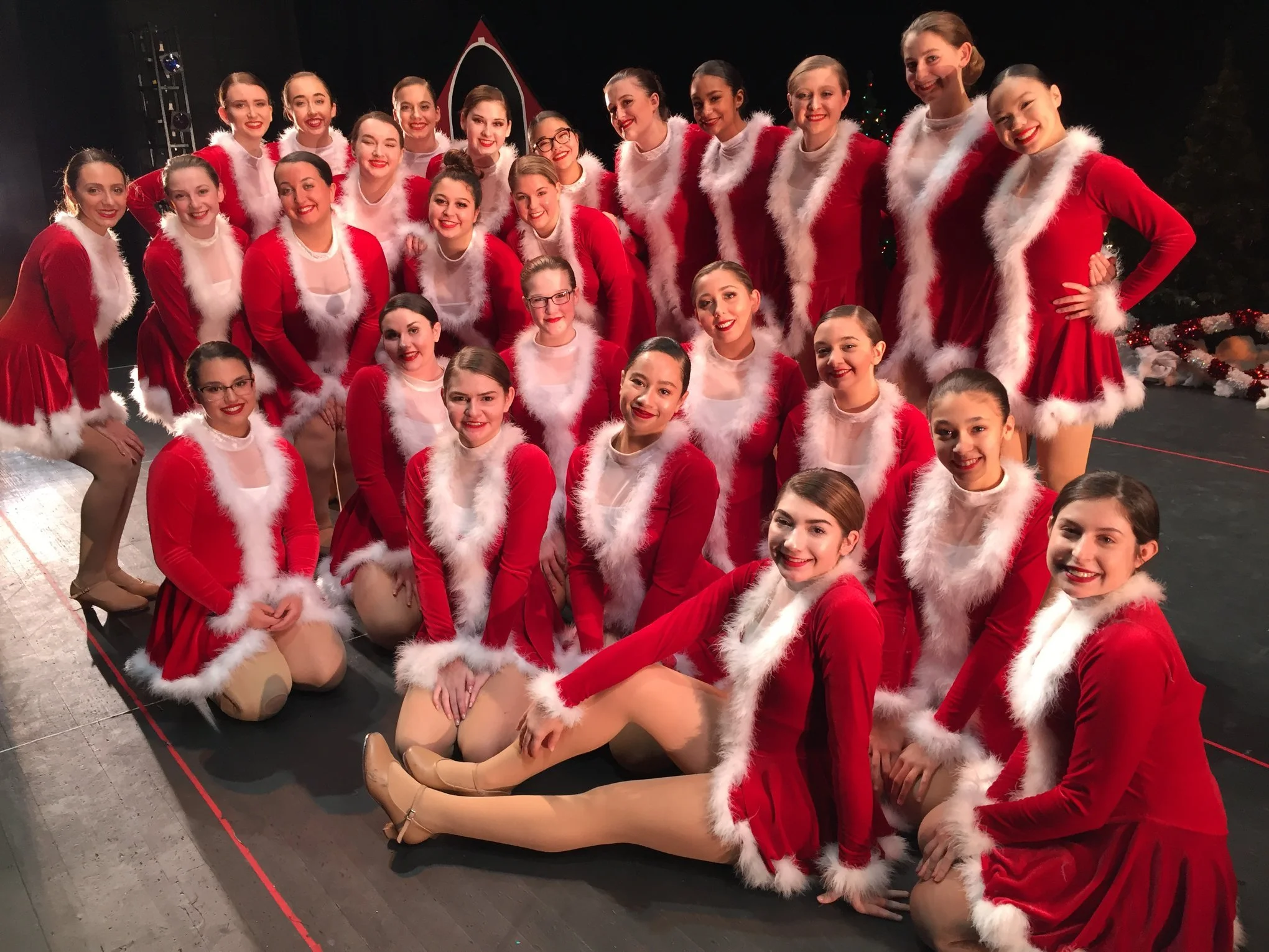 A group of young female performers dressed in red and white Christmas costumes, posing together on stage for a holiday photo.