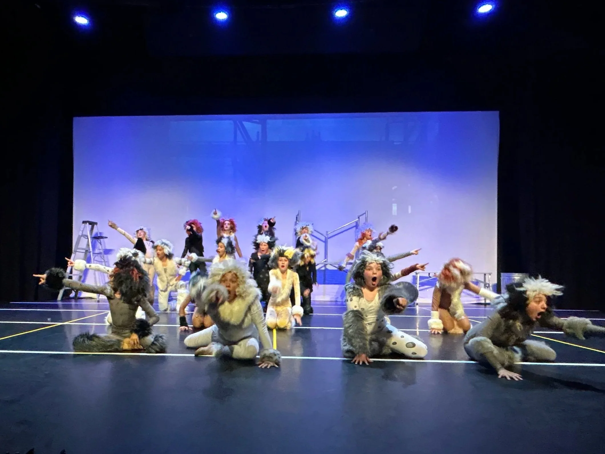 Theater cast in animal costumes performing on stage with a simple set, including a ladder and platform, under stage lighting.