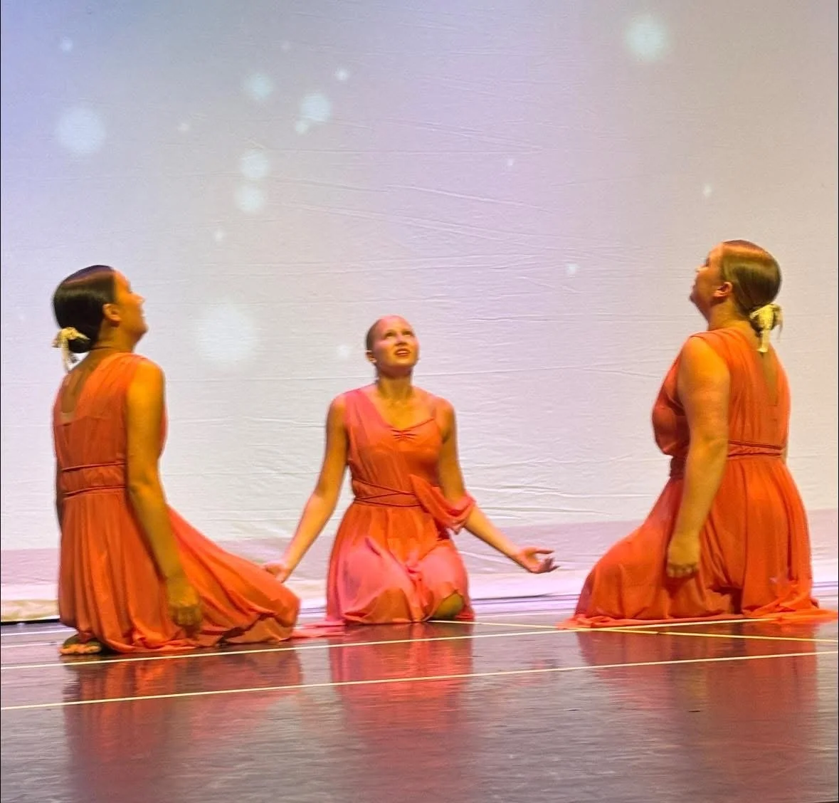 Three women in orange dresses kneeling on stage, looking upward, with a gray backdrop and star-like lights.