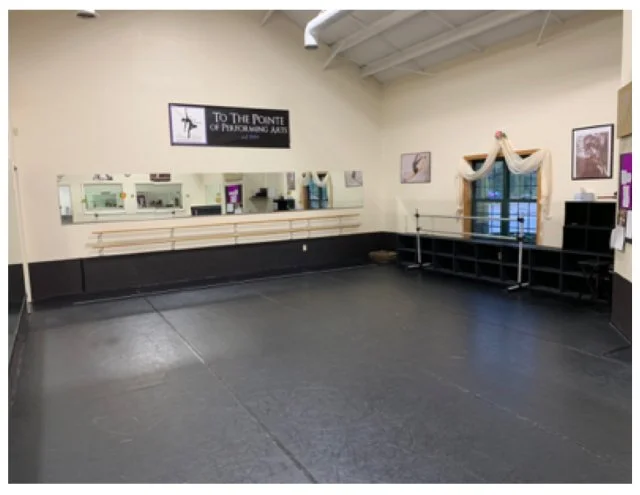 Empty dance studio with black floor, ballet barres, window with draped curtains, and religious signage on the wall.
