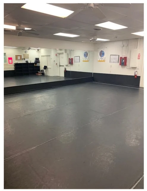 Empty dance studio with a large mirror, dark flooring, white walls, and ceiling lights.