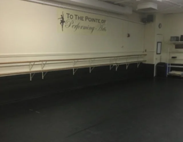 Empty dance studio with a ballet barre along the wall and a motivational sign reading "To the Pointe of Performing Arts".