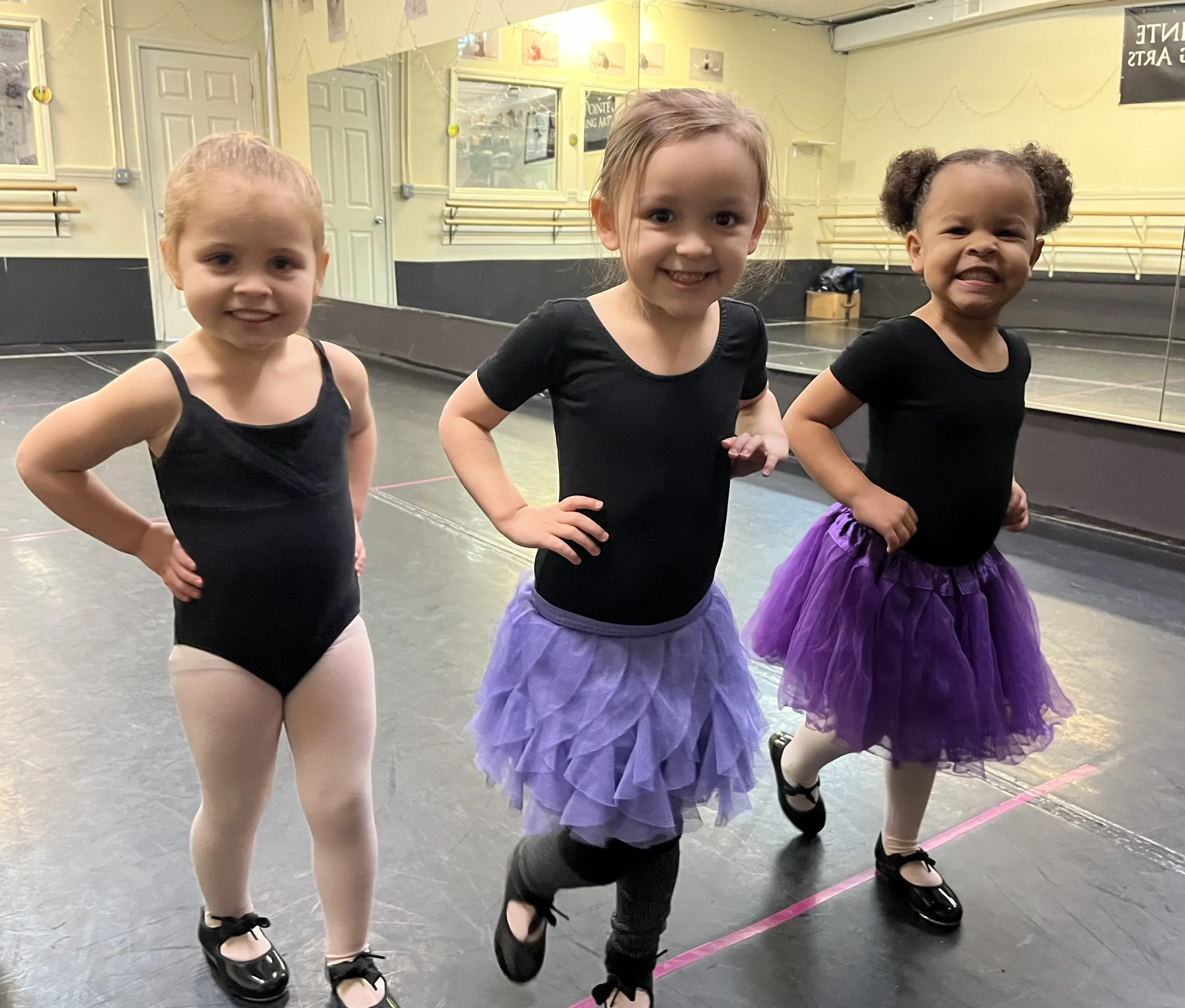 Three young girls in ballet outfits posing in a dance studio, smiling with hands on hips, wearing black leotards and purple tutus.