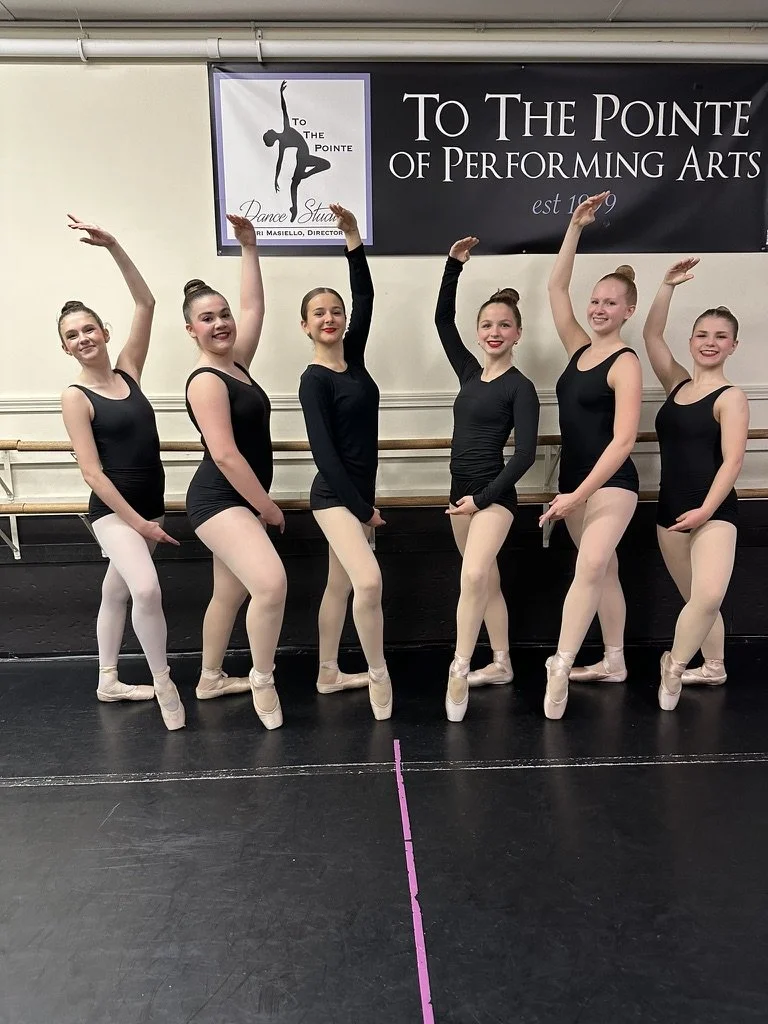 Six young ballet dancers in black leotards and pink tights standing on a ballet barre, posing with one arm raised, in a dance studio with a sign that reads "To the Pointe of Performing Arts."