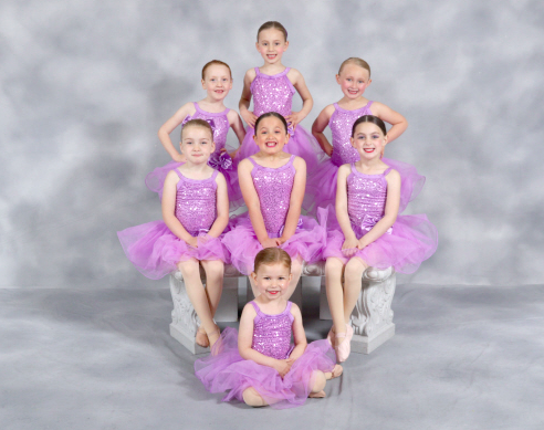 Seven young girls in purple ballet costumes posing together in a studio.