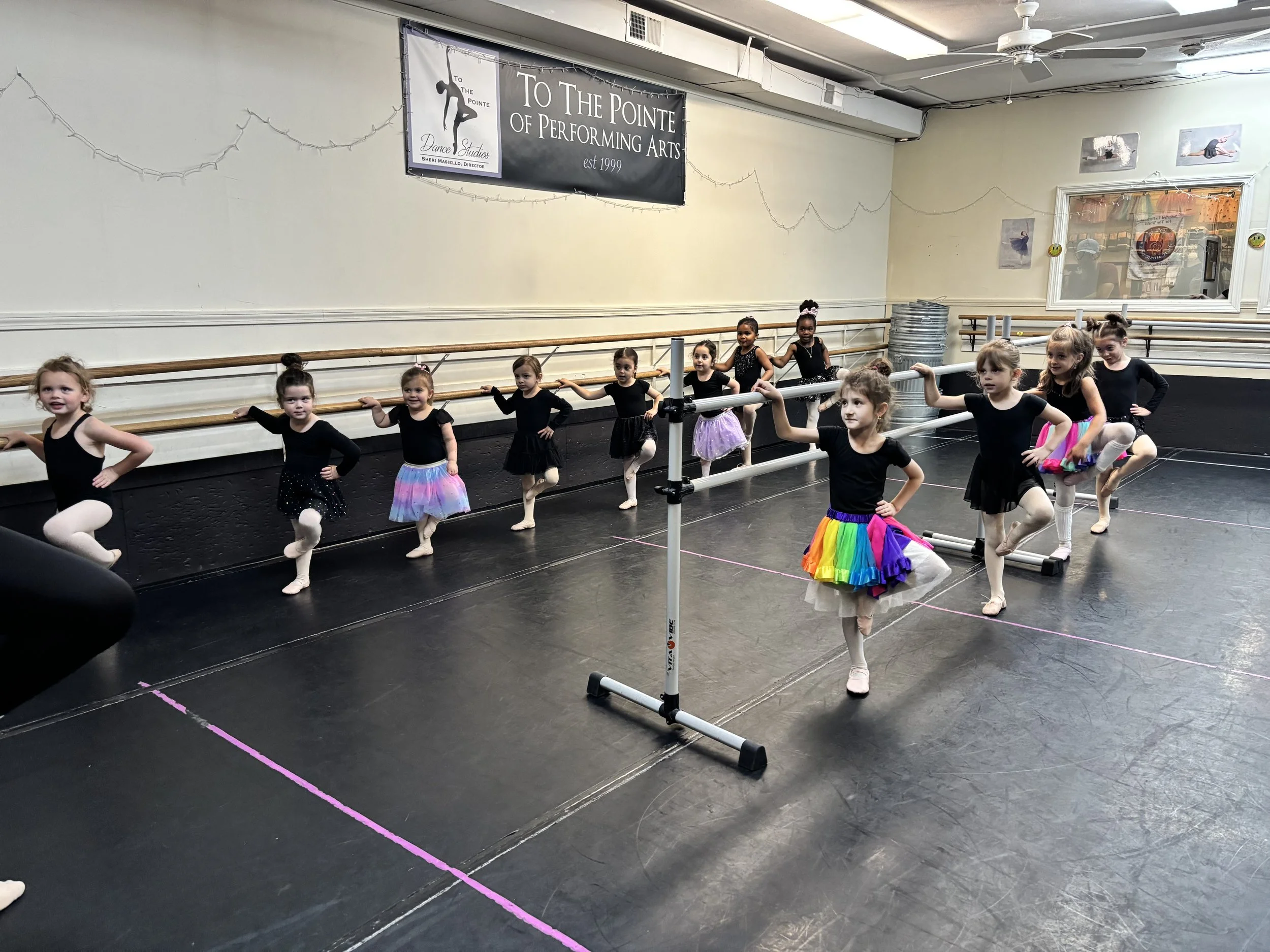 Young ballet students in black leotards and colorful tutus practicing at a dance studio with mirrors and ballet barres.