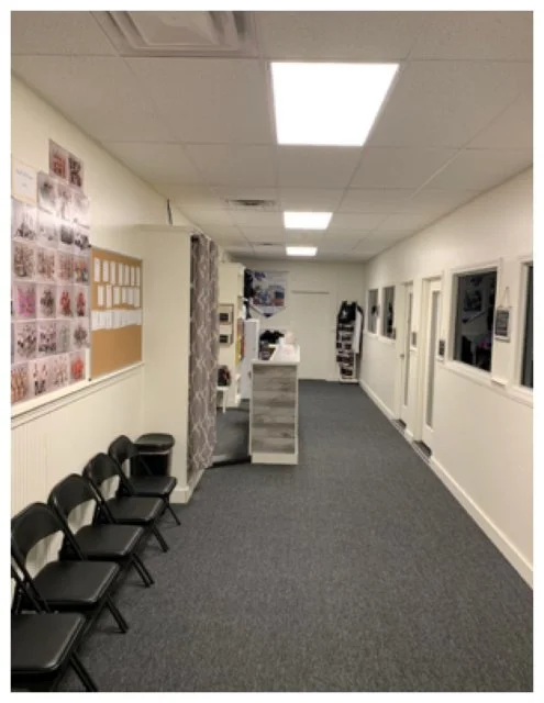 Empty waiting area with black chairs along a wall, bulletin board, and reception desk in a hallway.
