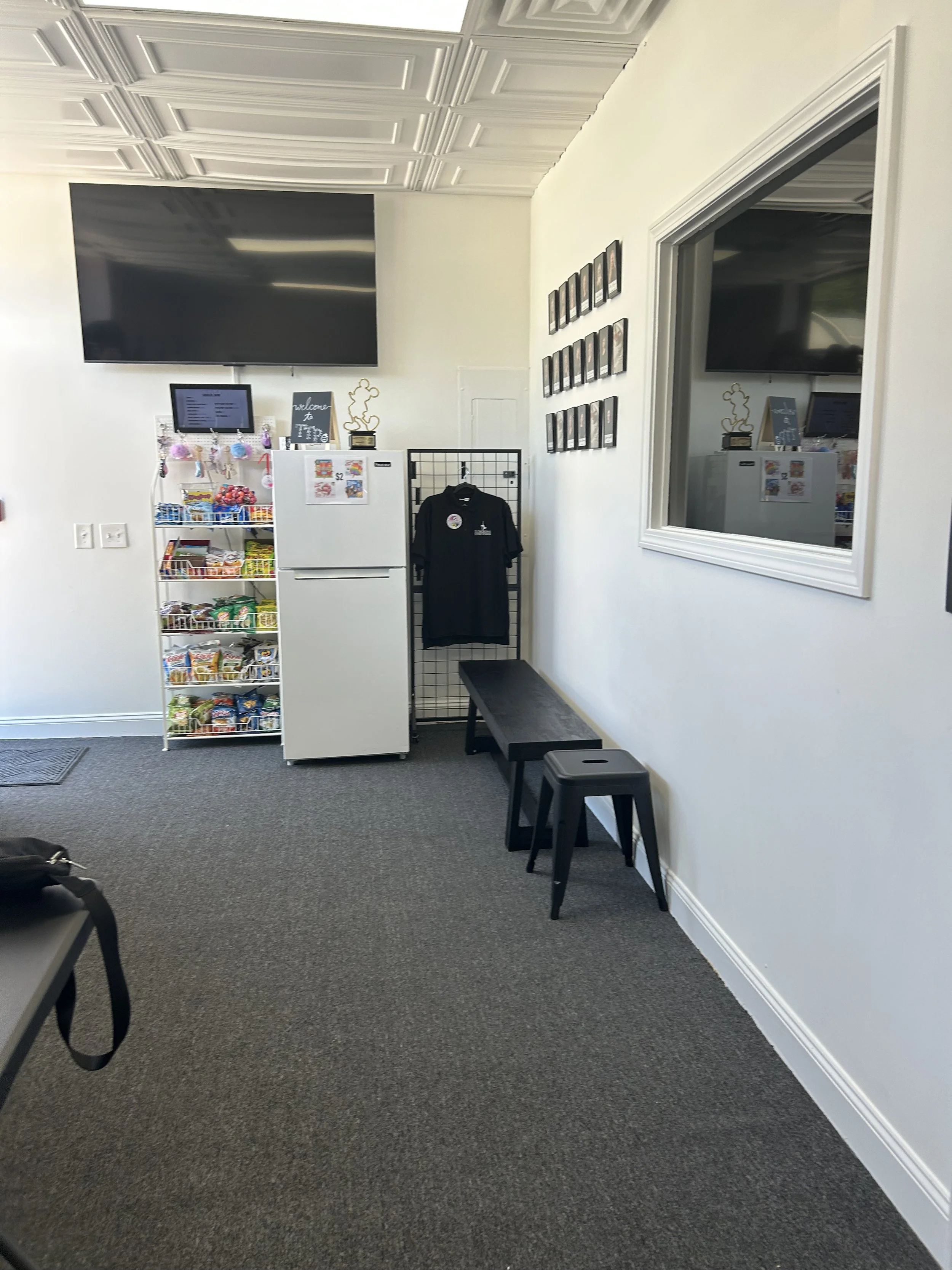 An interior view of a room with white walls and black furniture, including a black bench, black stools, and a black shirt hanging on a black wire grid. There is a white refrigerator with snacks and a small chalkboard sign on top, a small screen displaying text, and multiple framed photos on the wall. A large flat-screen TV is mounted on the wall, and a window reflects part of the room.