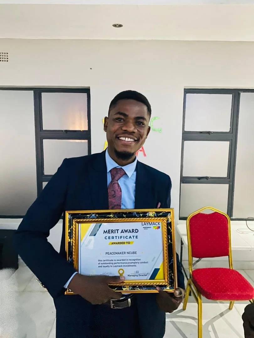 A woman stands in front of a painted brick wall with a sign for Zara Center Trust. She is wearing a black t-shirt with a graphic design, blue jeans, and flip-flops, looking slightly to her right with her hands behind her back.