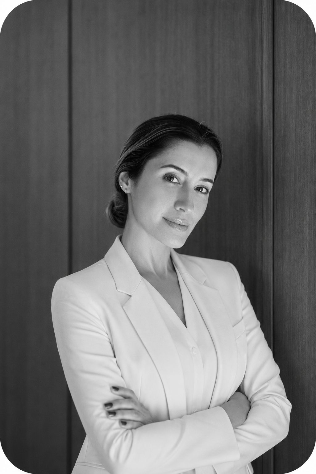 Mulher jovem de cabelo preso, usando blazer claro, com braços cruzados, posando de lado contra fundo de madeira, expressão confiante, em foto preto e branco.