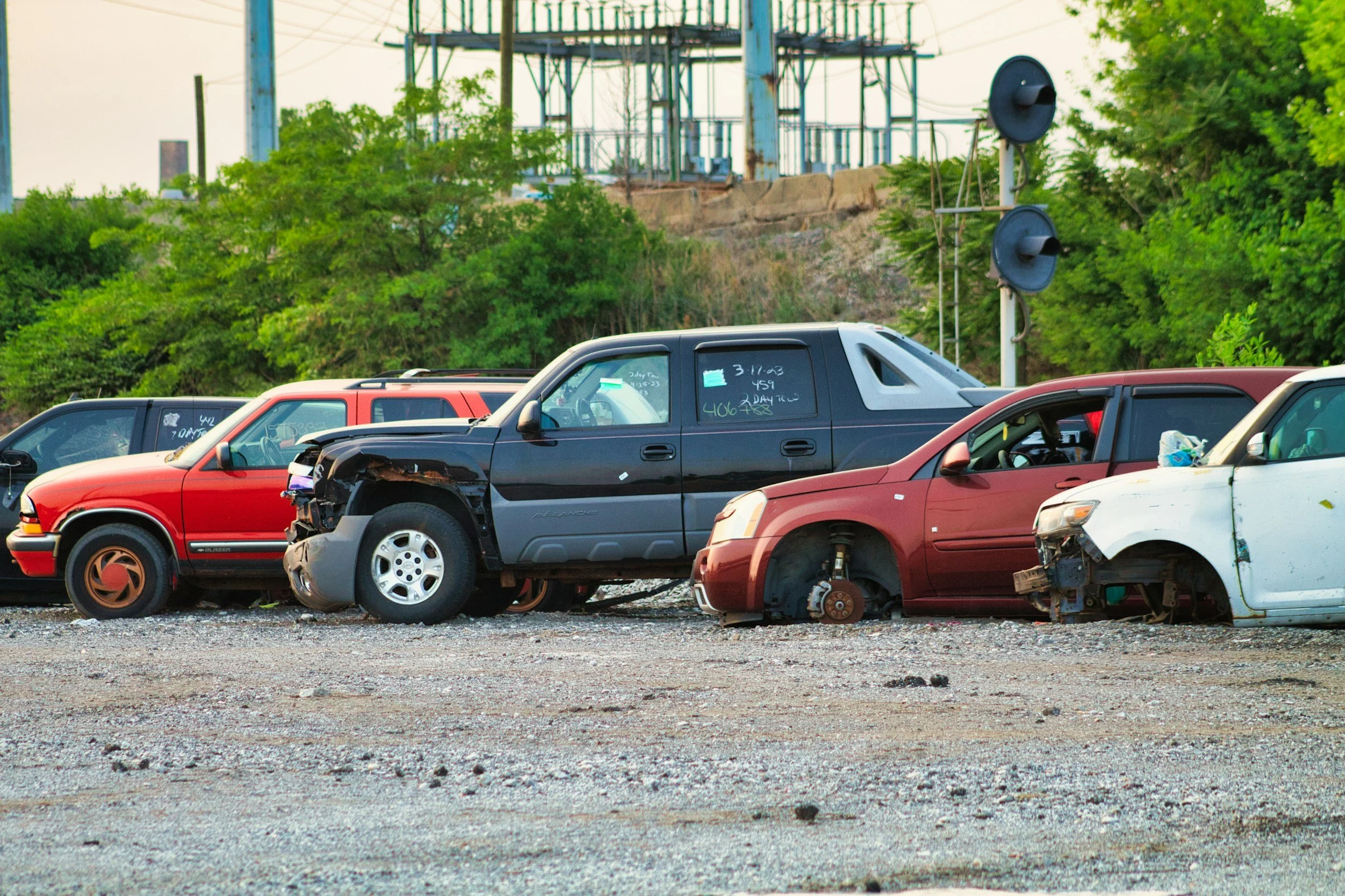 A row of damaged and wrecked cars parked outdoors, some missing wheels, with a background of green trees and industrial power lines.