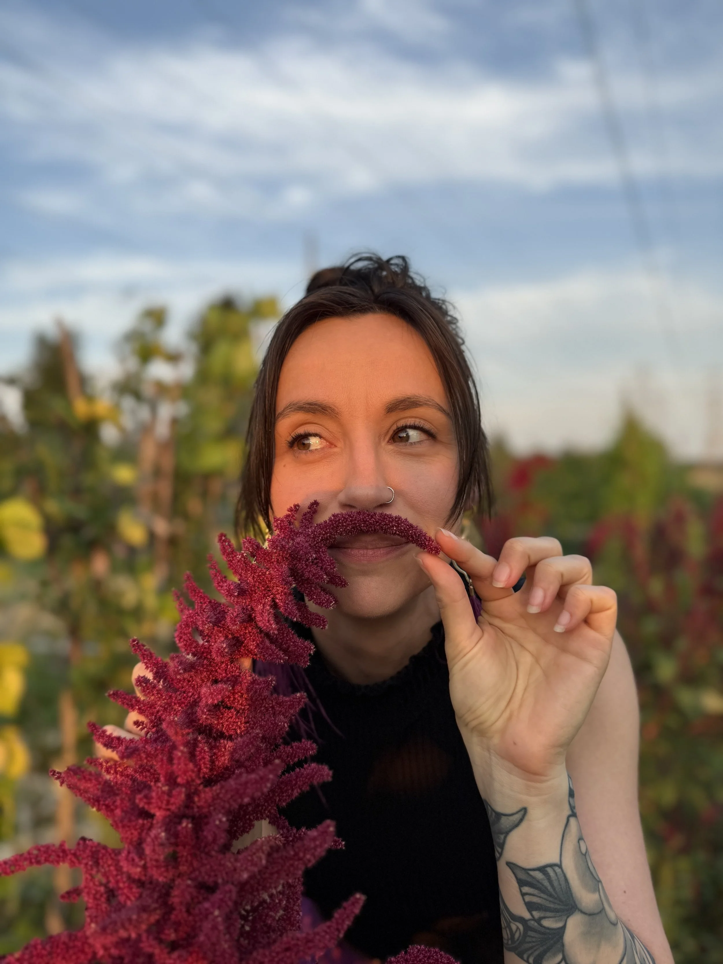 A femme person with dark hair and a nose ring playfully posing with a red flower under her nose as mustache, standing outdoors in a garden during sunset.