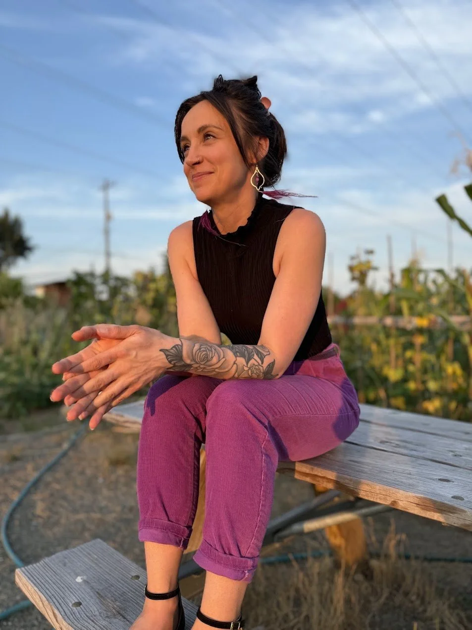 A femme person with dark hair and a nose piercing sitting on a wooden bench outdoors during sunset, wearing a black sleeveless top, purple pants, and black sandals, with a tattoo of roses on her arm, smiling and looking away.