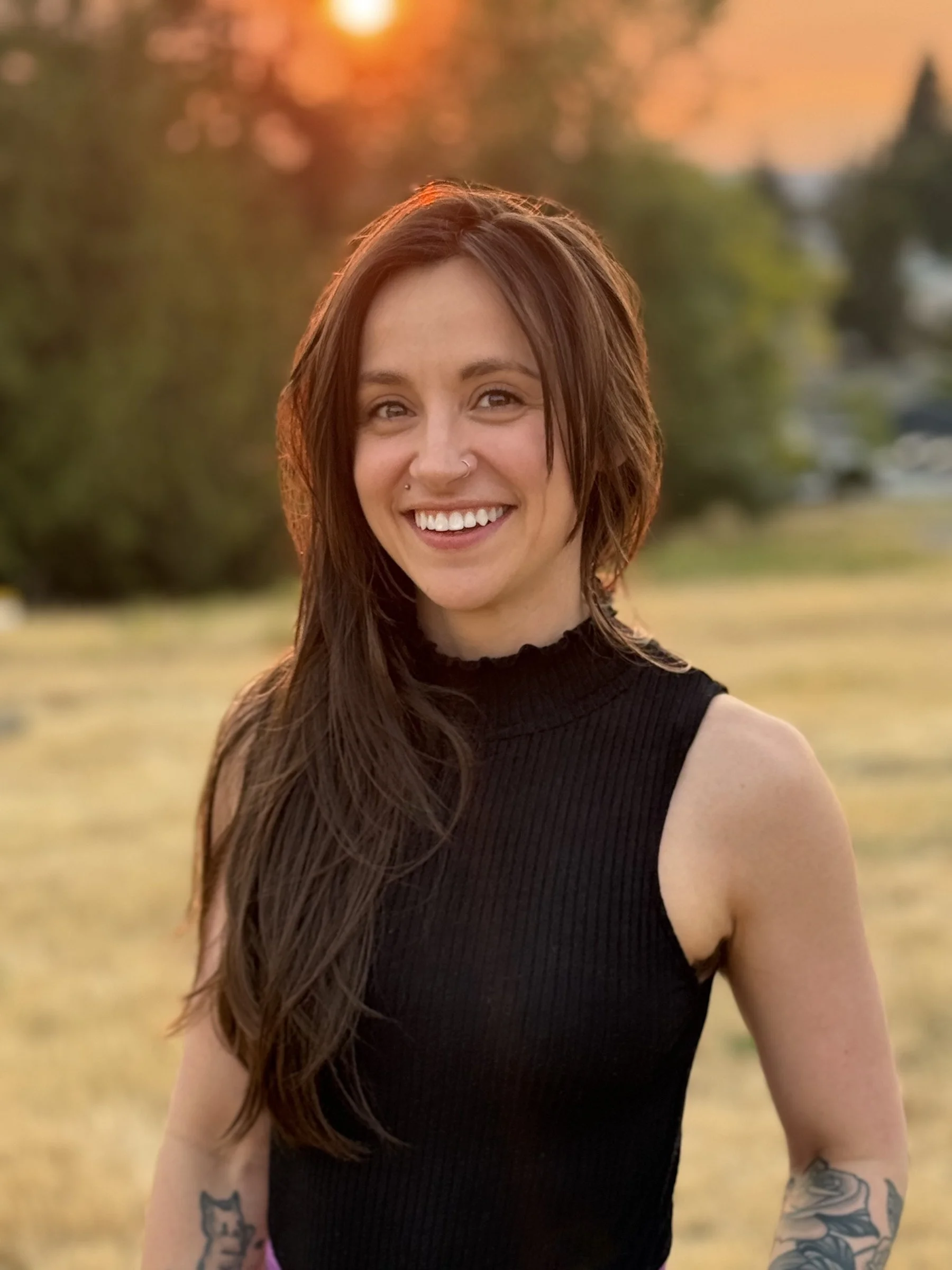 A smiling femme person with long brown hair and tattoos on her arms, wearing a sleeveless black top, outdoors during sunset.