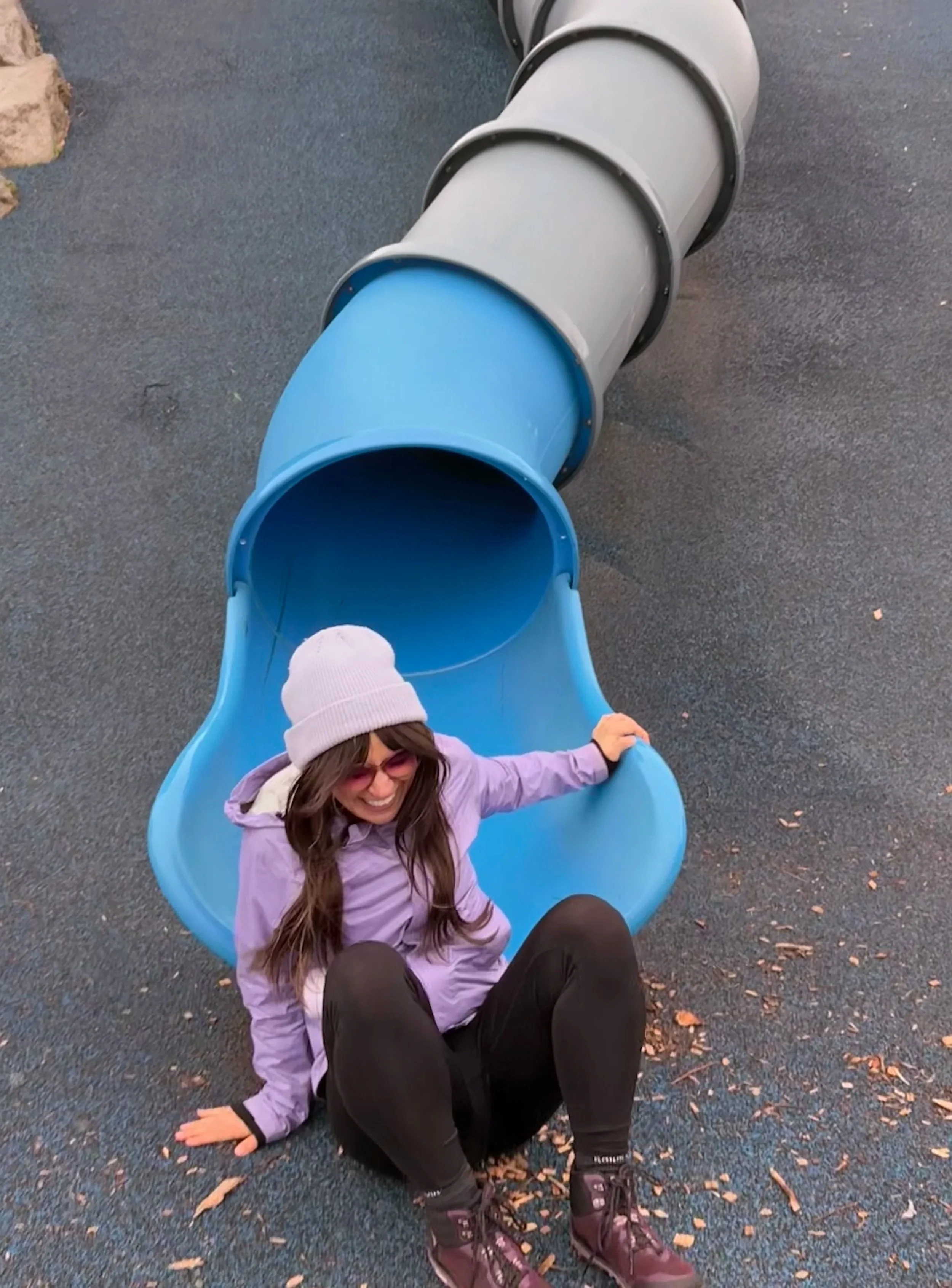 A smiling person with long hair, wearing a white beanie and purple jacket, sitting at the bottom of a blue slide in a playground, on a blue rubber ground surface with small debris.