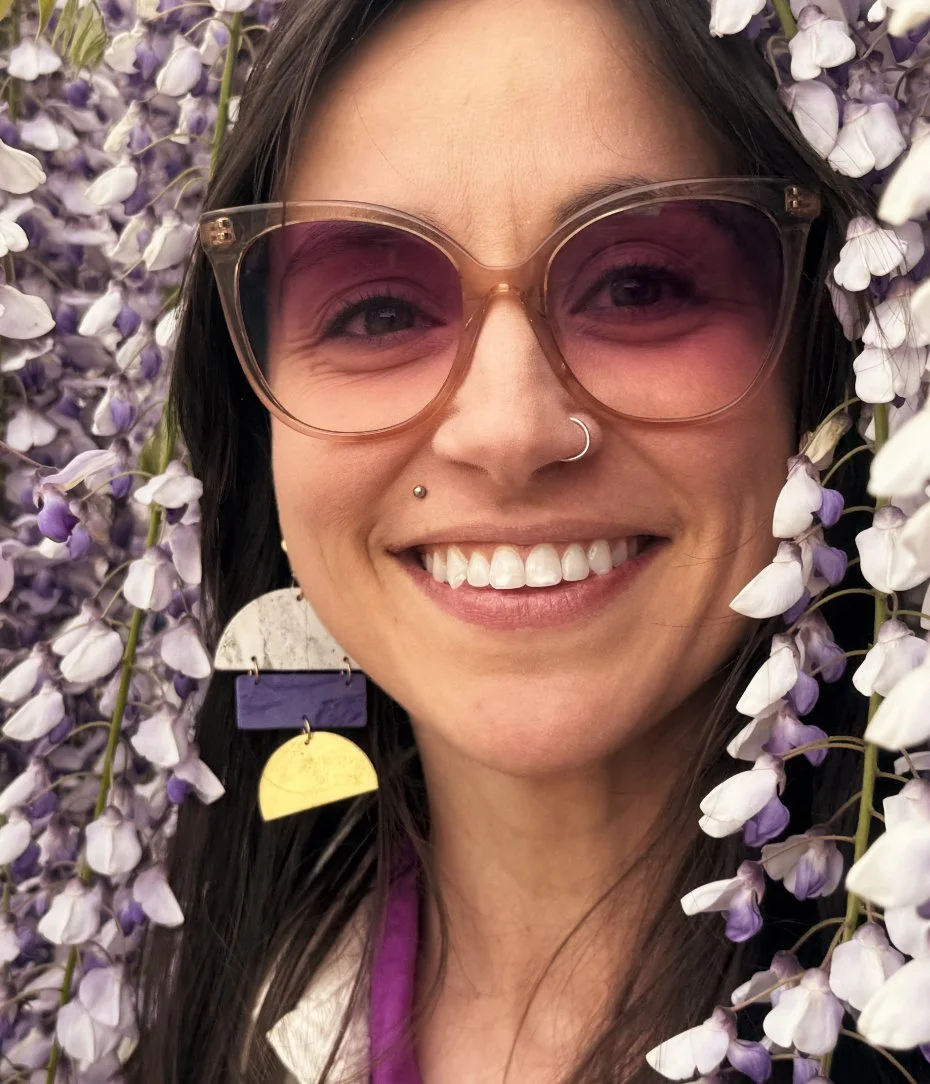 A femme person smiling with lilac gradient sunglasses, surrounded by purple and white flowers.