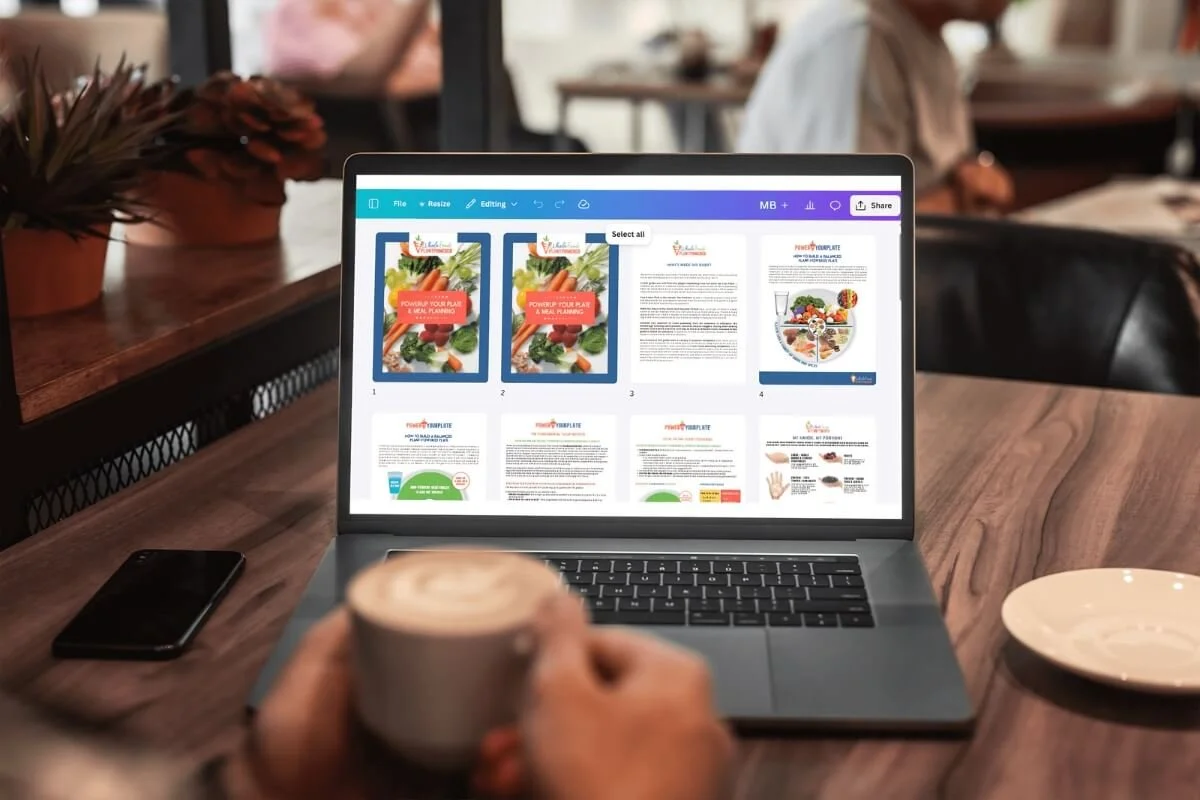 A person in a cafe working on a laptop displaying a presentation or brochure about nutrition or wellness, with a cup of coffee or tea in hand, a smartphone on the table, and a plate nearby.