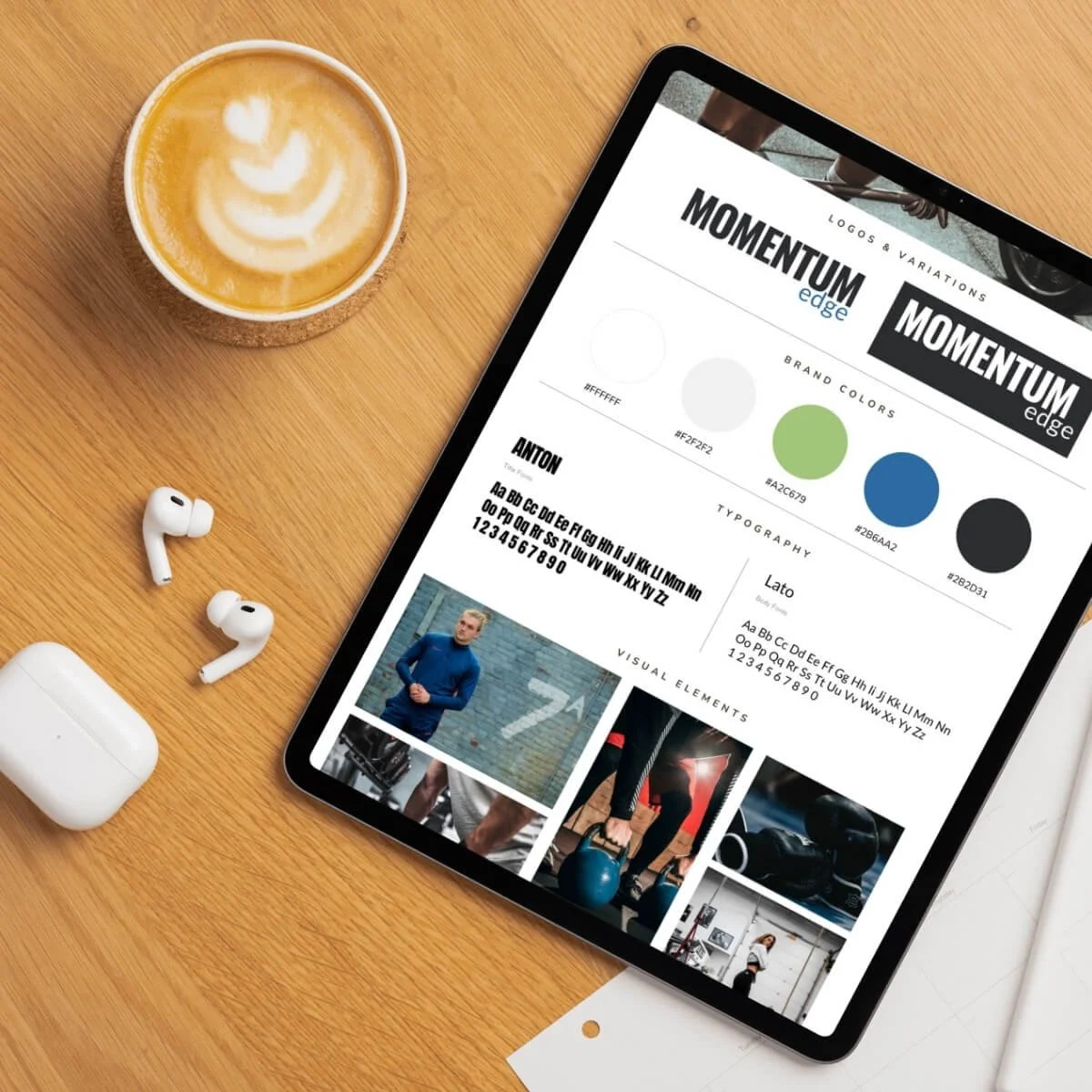 A wooden desk with a cup of coffee, wireless earbuds with a case, and a tablet displaying a design board for "Momentum Edge" brand with color palettes, fonts, and images.