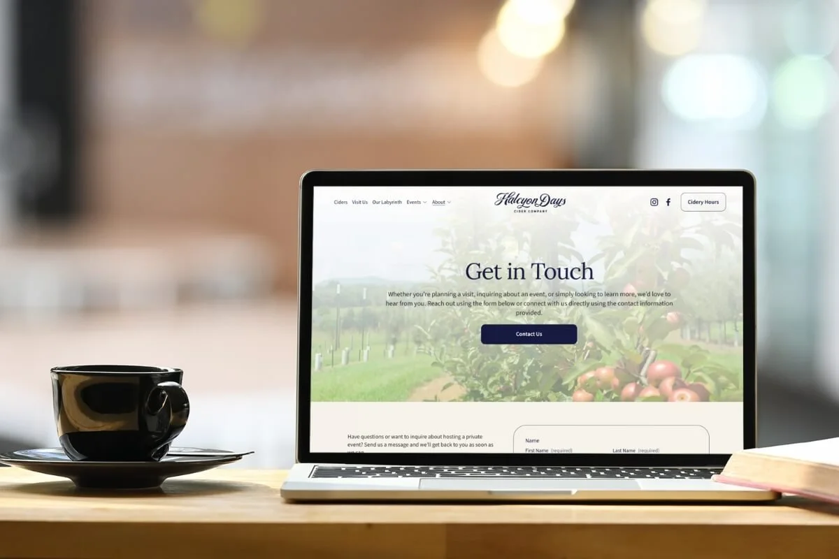 A laptop displaying the 'Get in Touch' page for Halcyon Days Cider Company, with a black coffee cup on a saucer beside it, on a wooden desk.