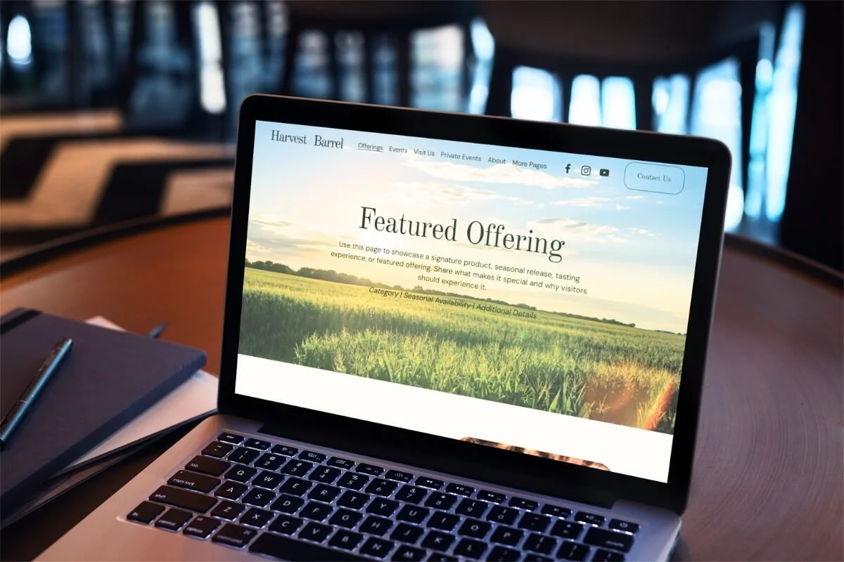 Laptop on a wooden table displaying a website titled 'Harvest Barrel' with a feature about seasonal product offerings, showing a scenic image of a green field and sky in the background.