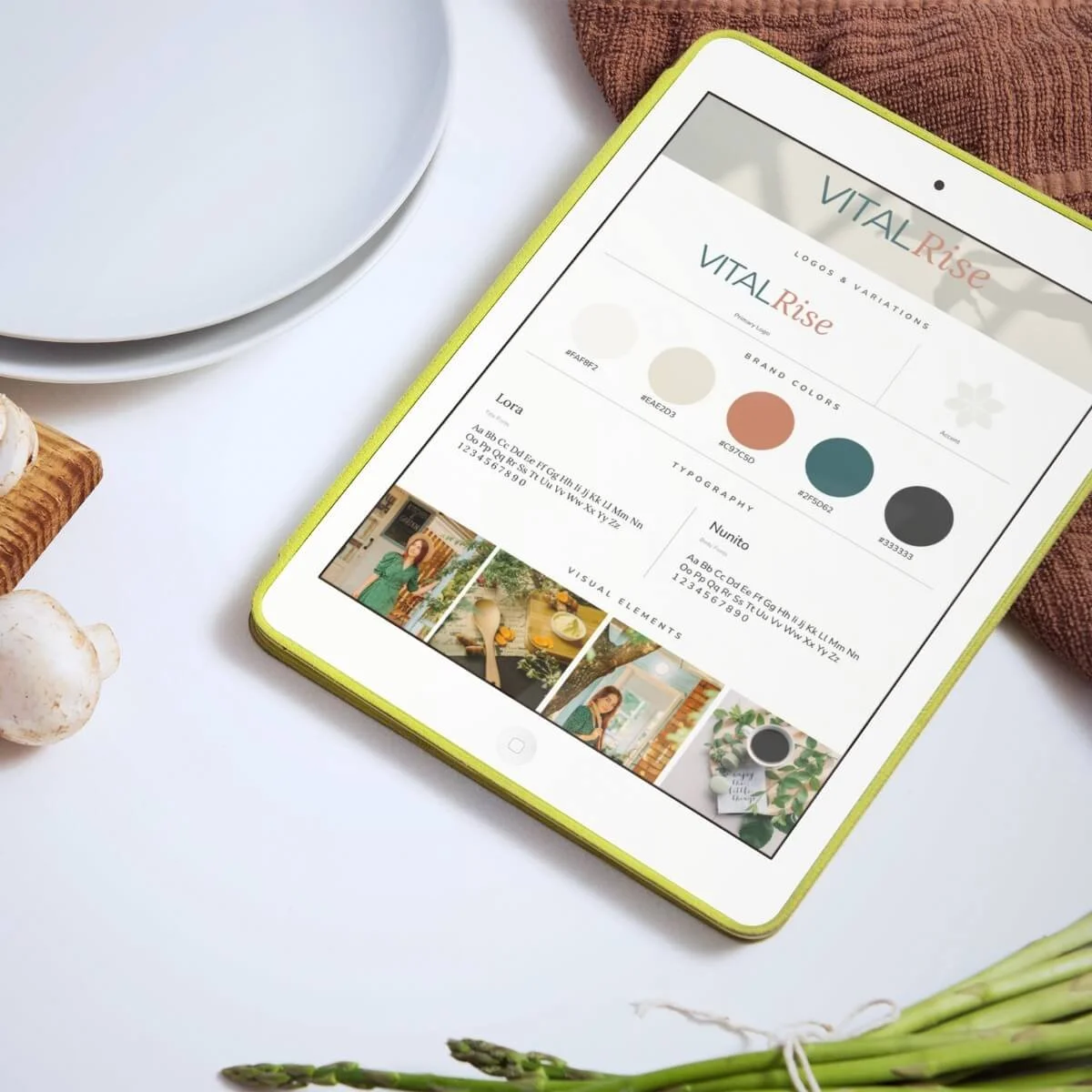 Tablet displaying a branding design project titled 'VITAL Rise' with color palettes, typography, and visual elements. Surrounding objects include white bowls, a brown towel, garlic, and asparagus on a white surface.