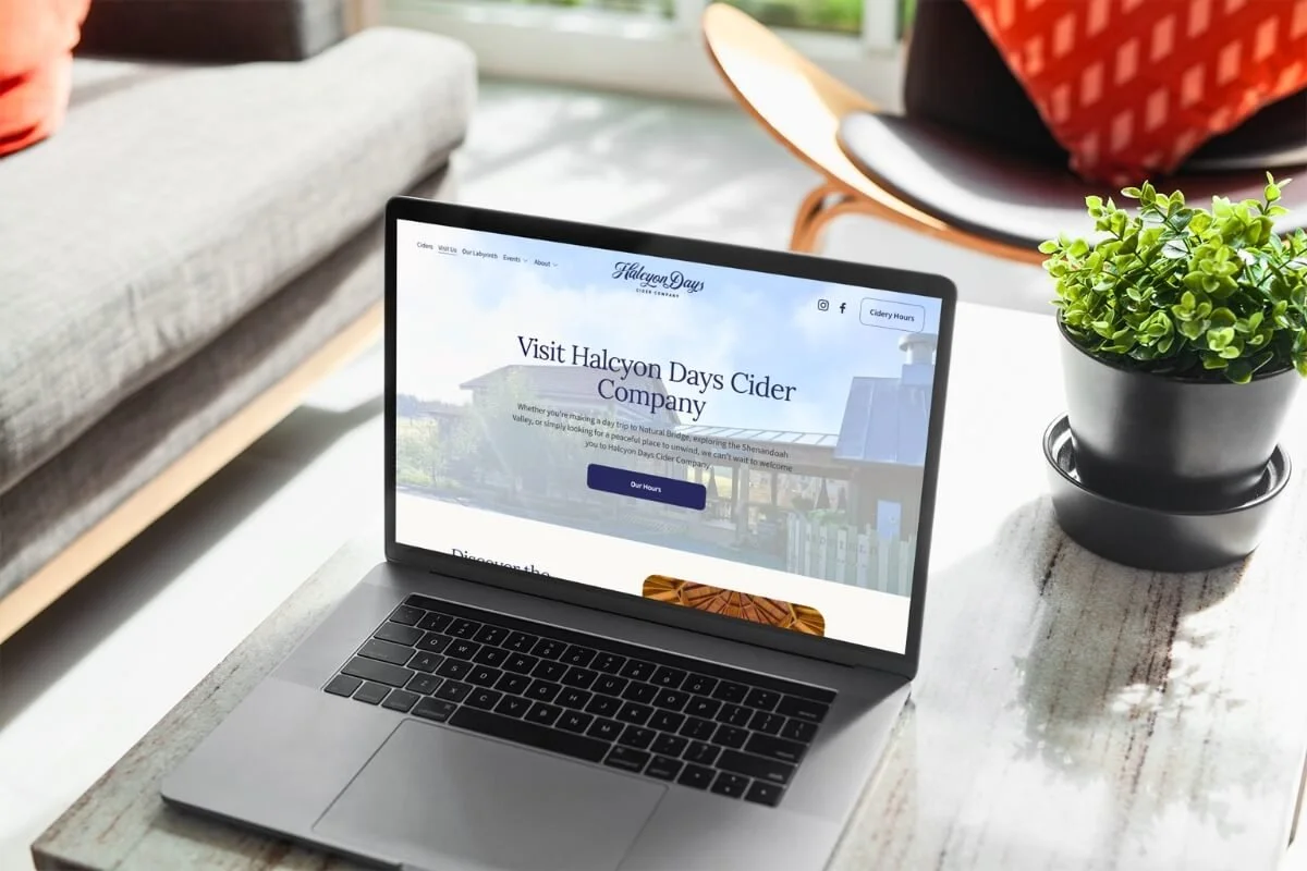 Open laptop on a wooden table displaying the Halcyon Days Cider Company website, next to a black pot with a green plant, in a cozy indoor setting with a gray couch and an orange chair in the background.