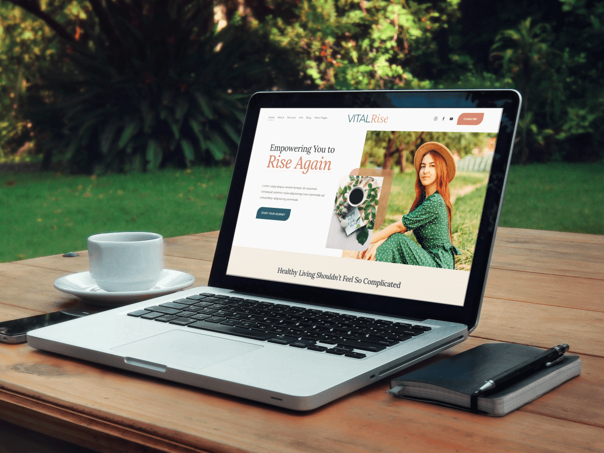 A laptop on a wooden outdoor table displaying a website titled "VITAL Rise" with a woman in a green dress and straw hat. Next to the laptop is a cup on a saucer, a closed black notebook with a pen on top, and a smartphone in a case. The background features green grass and trees.