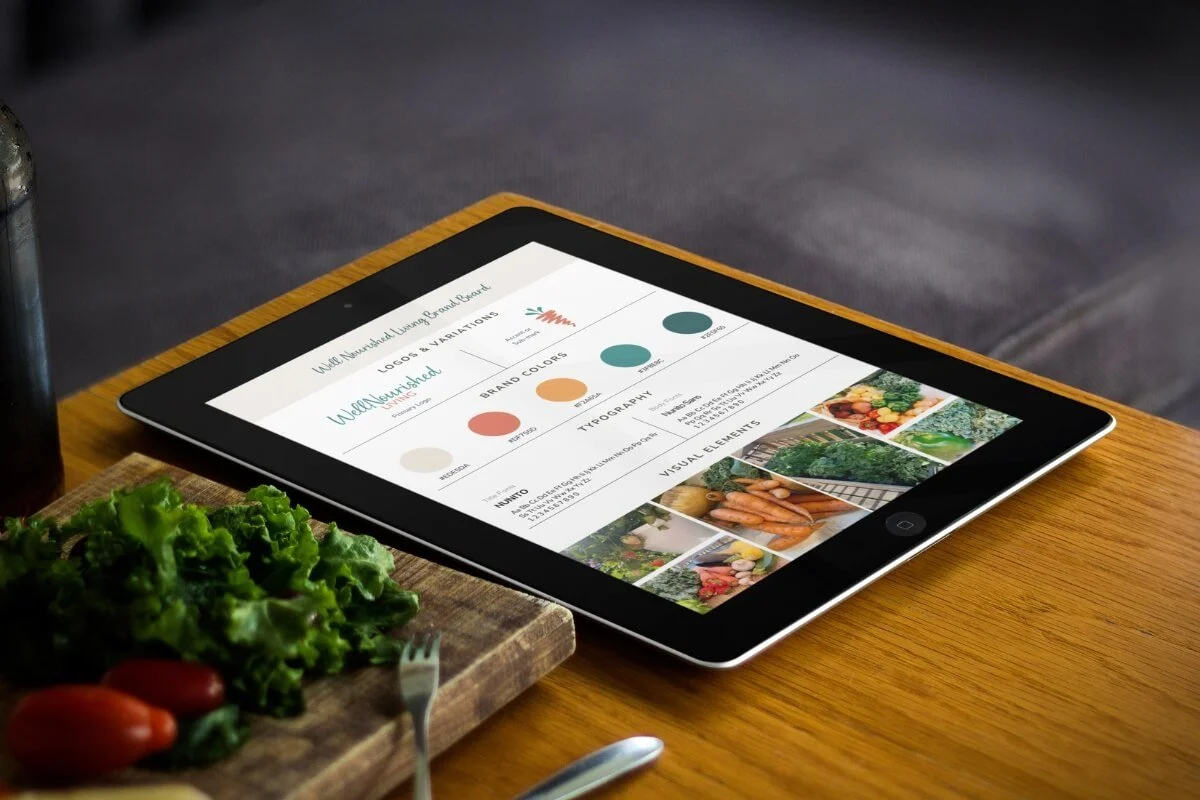 Tablet displaying a brand style guide on a wooden table with a plate of salad and tomatoes nearby.