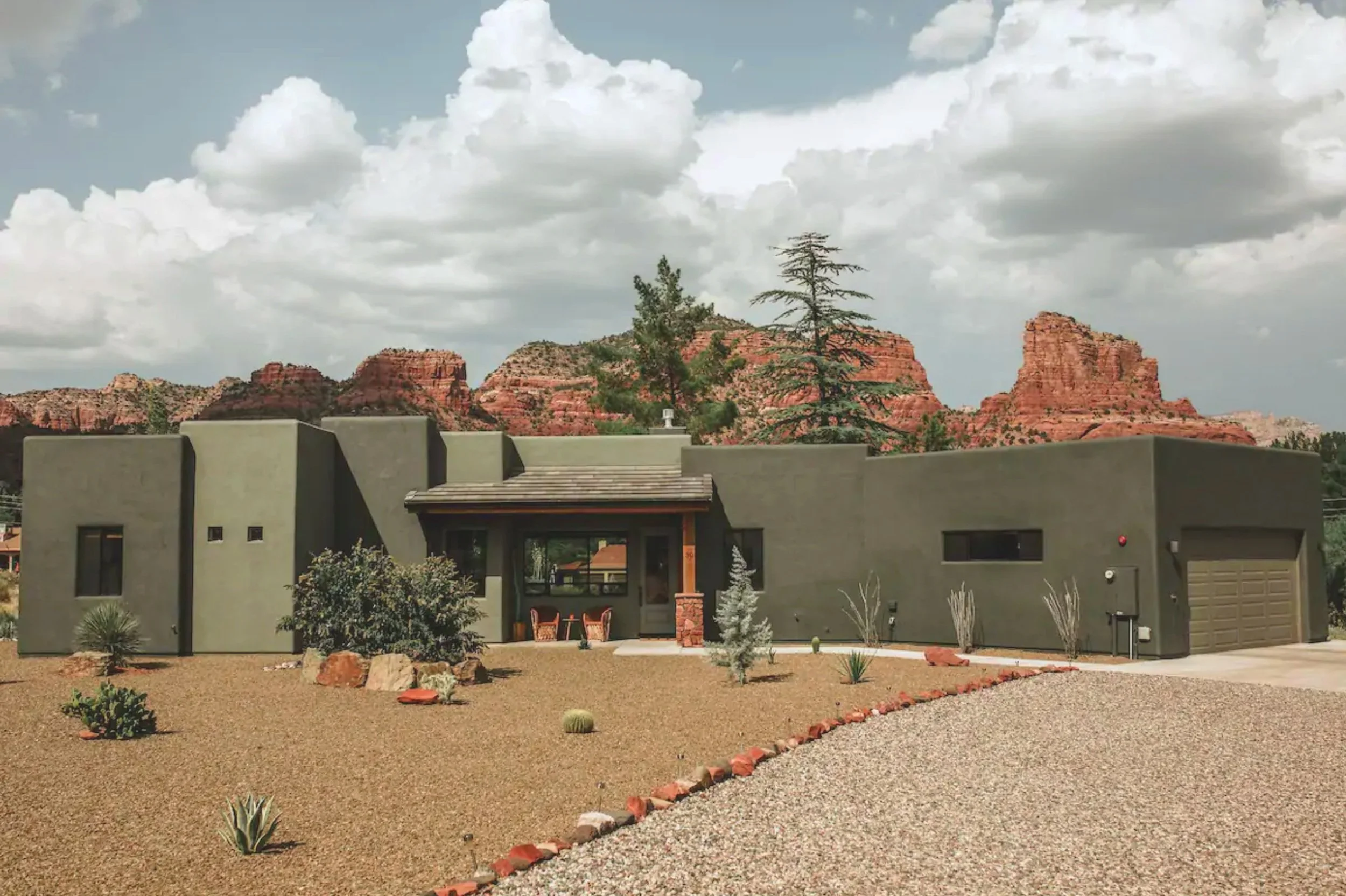 A modern house with a desert landscape in front, featuring various cacti and rocks, set against a mountainous backdrop with red rock formations and a partly cloudy sky.