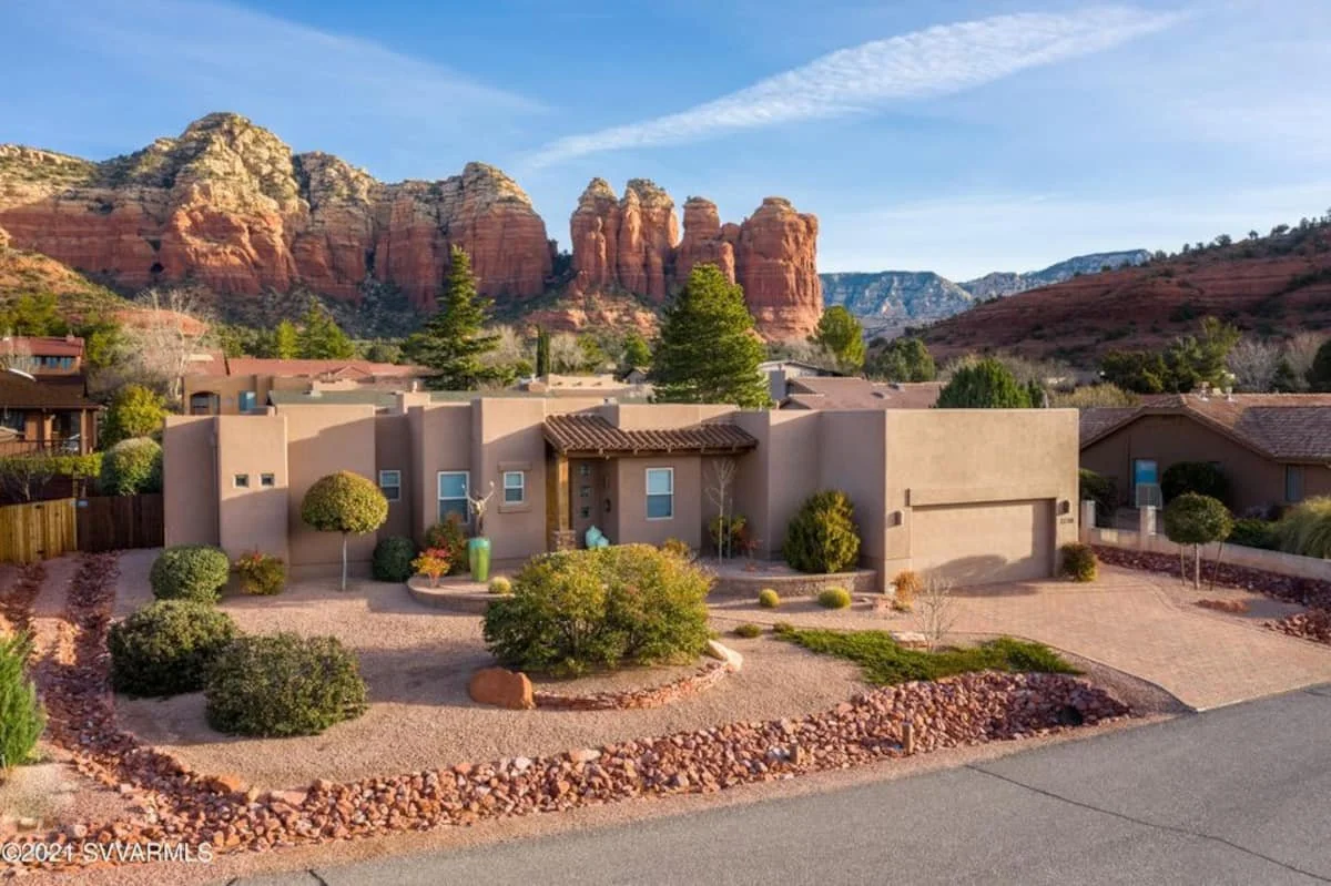 A house with a desert landscape in front, featuring gravel, rocks, and desert plants, with red rock formations and mountains in the background under a blue sky.